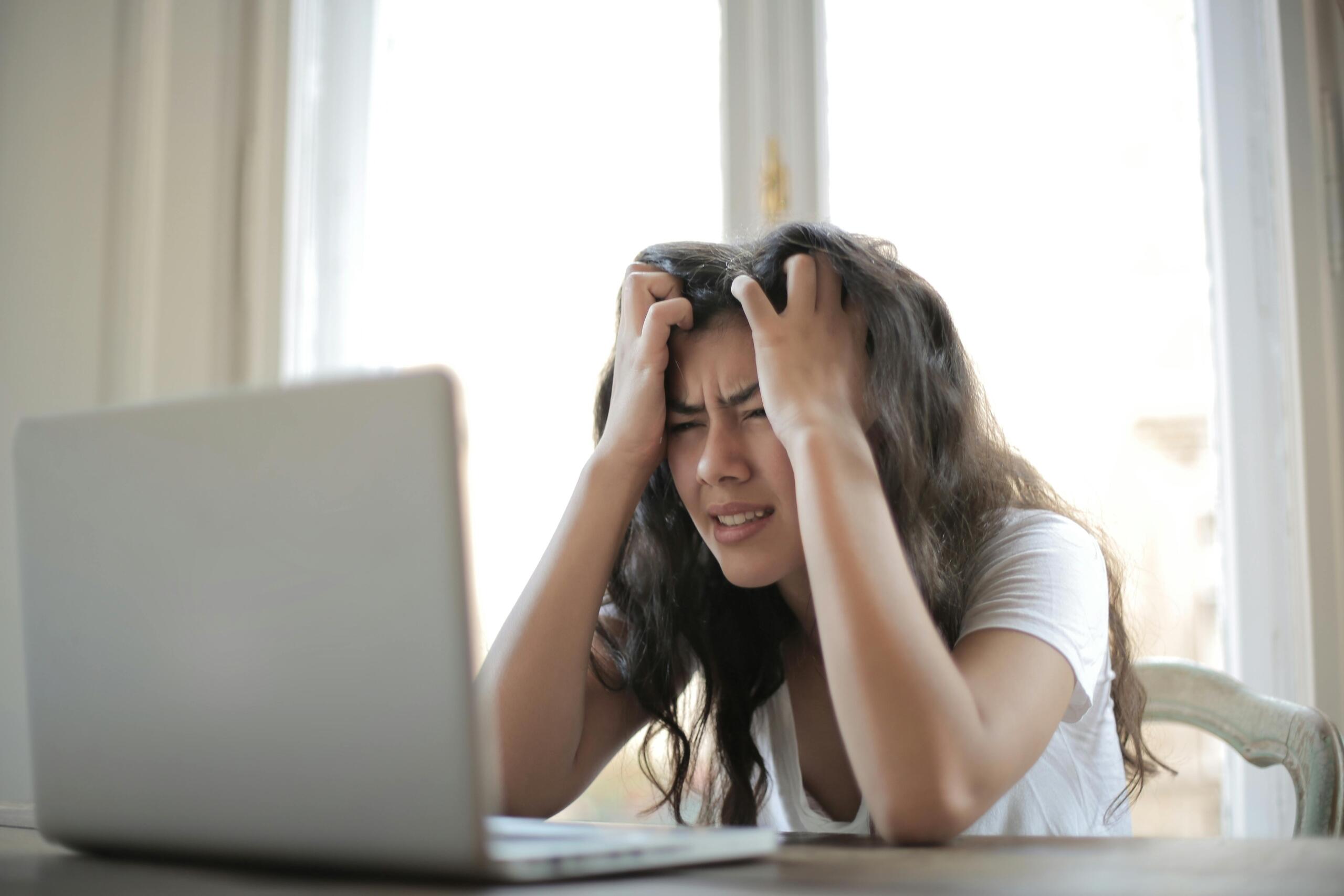 A woman sits at a desk with a laptop, hands on her head, expressing frustration or stress in a sunlit room.