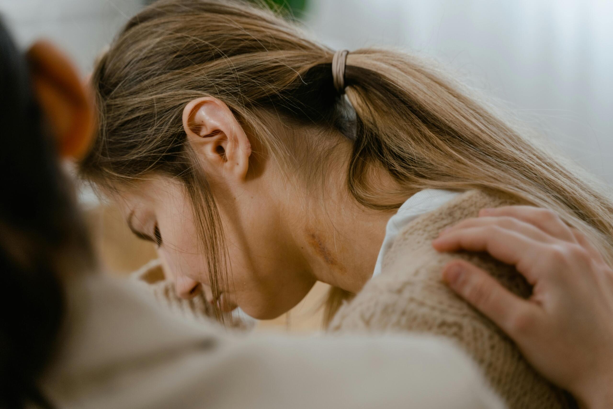 A person with long hair is shown from behind, gently comforted by another, wearing a cozy sweater in a softly lit room.