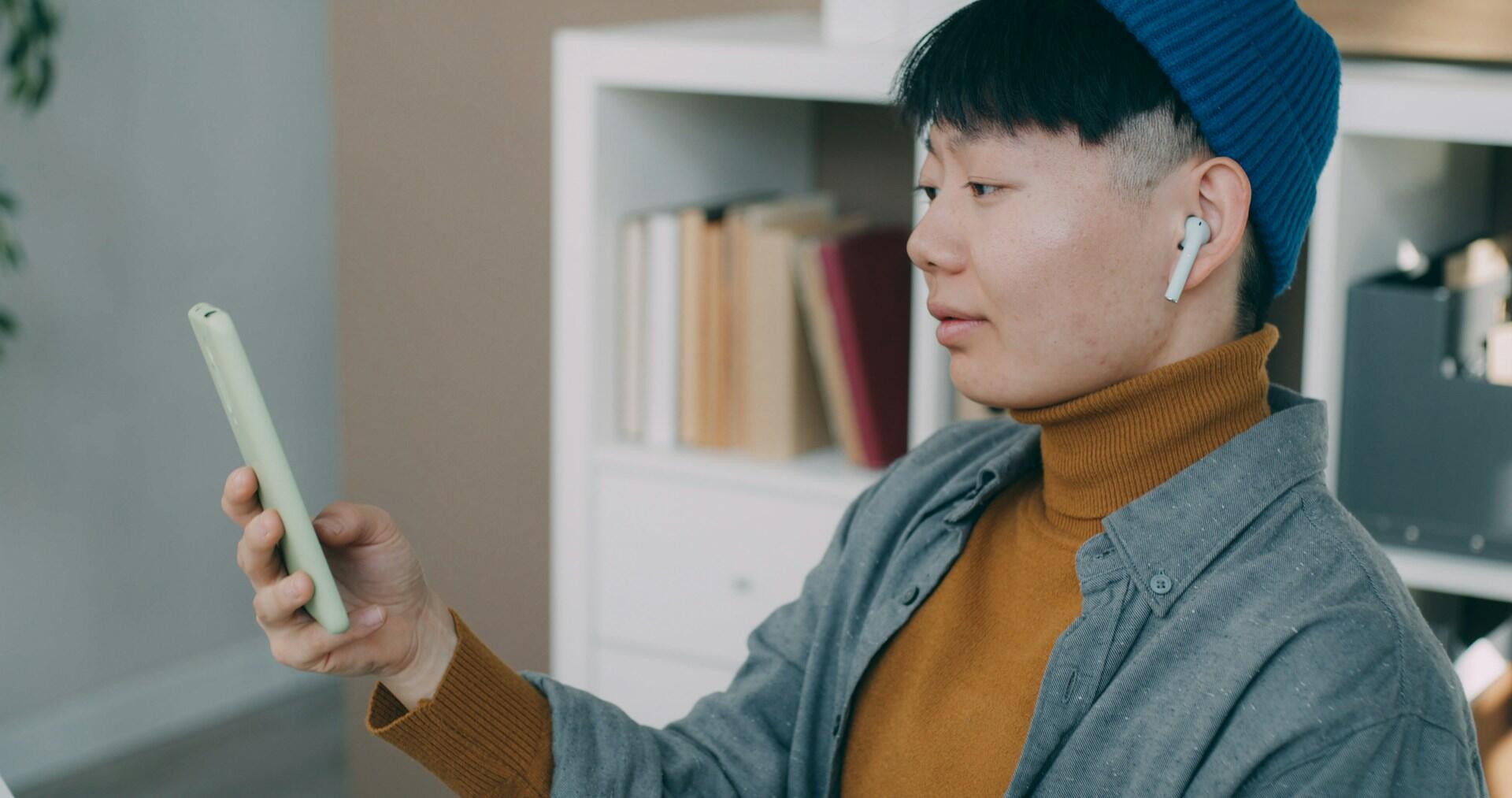 A person in a blue beanie and gray shirt holds a smartphone, wearing wireless earbuds, in a cozy, modern workspace.