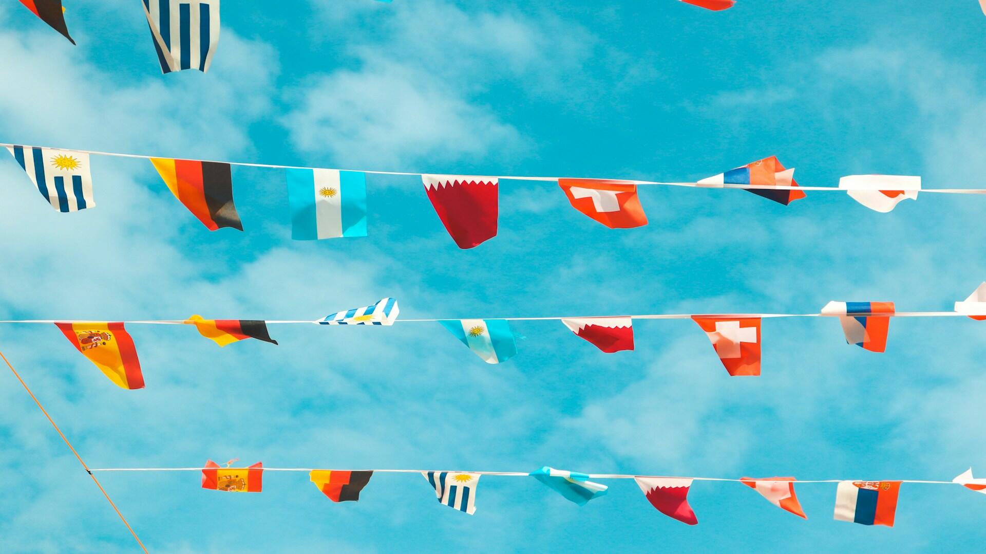 Colorful international flags hang between thin lines under a bright blue sky with fluffy clouds, celebrating global unity and diversity.