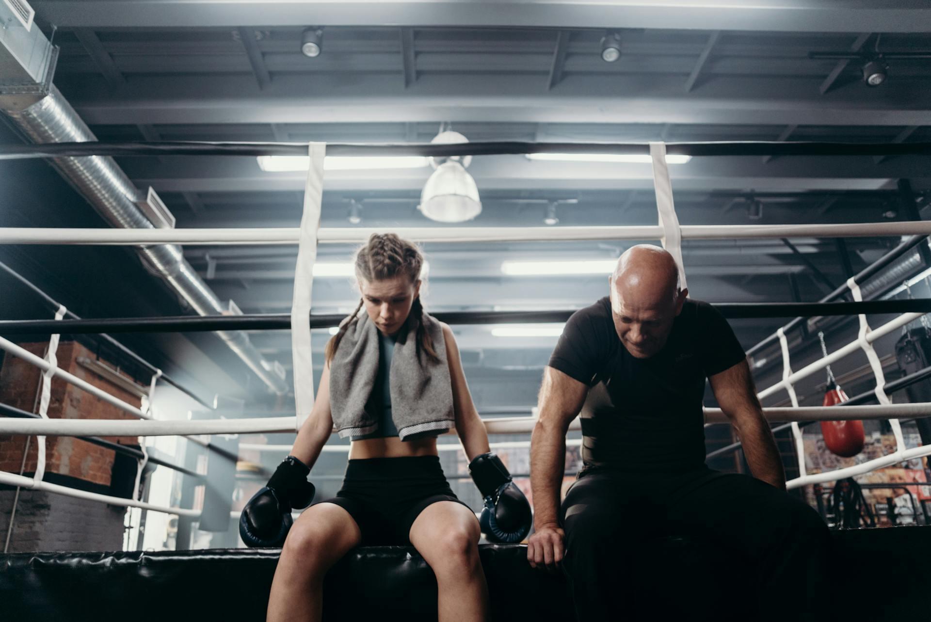 A boxer in workout gear sits on a gym ring's edge, resting with gloves on, while a trainer sits nearby, focused and engaged.