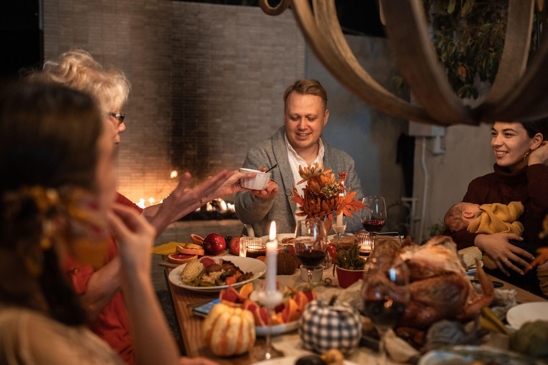 A family gathers around a candlelit dinner table with a roast turkey; a man passes a bowl while a woman holds a baby.