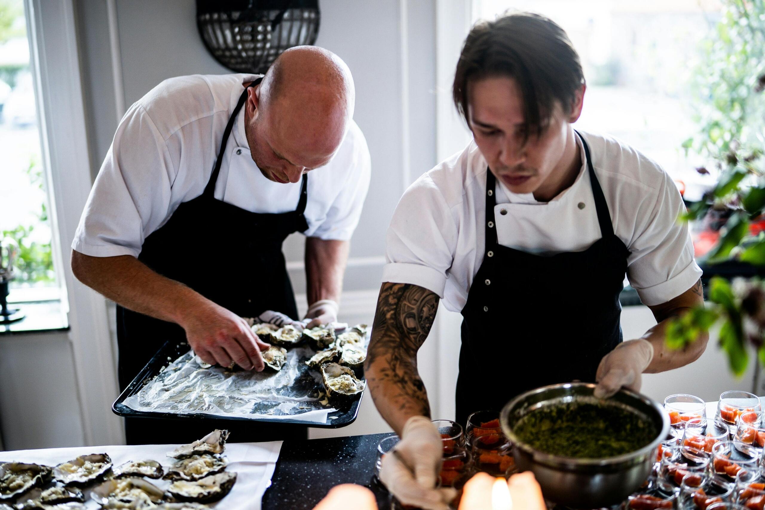 Two chefs work side by side in a kitchen, focused on preparing dishes at their stations as steam rises around them.