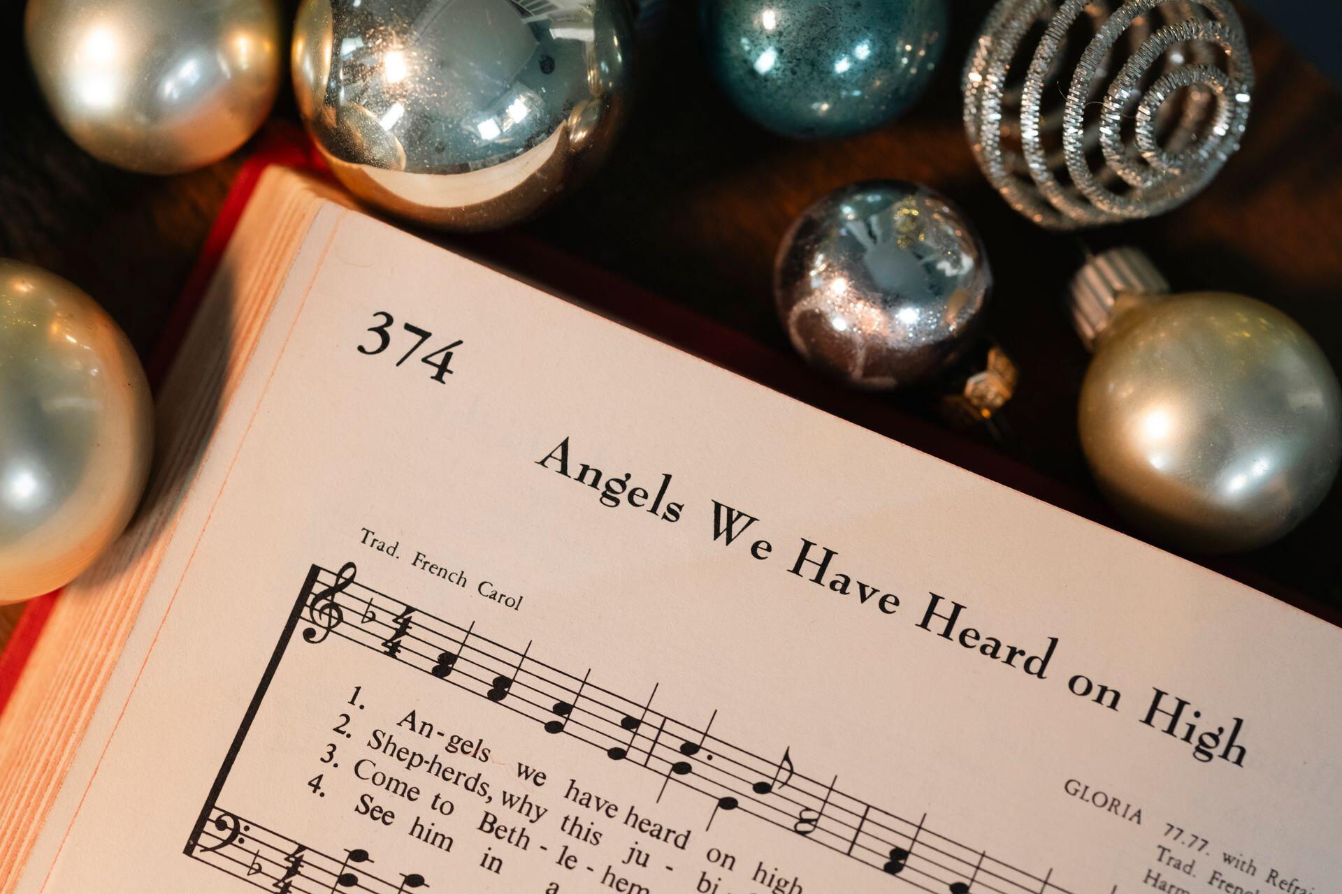 An open hymnal showing page 374, “Angels We Have Heard on High,” with printed sheet music, surrounded by silver Christmas ornaments.