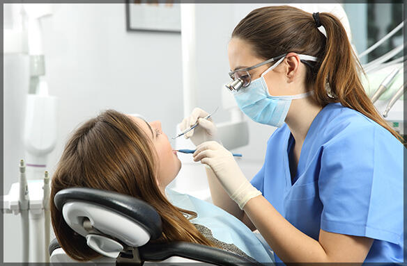A dental hygienist in a blue uniform is using instruments to clean a patient's teeth in a modern dental office.