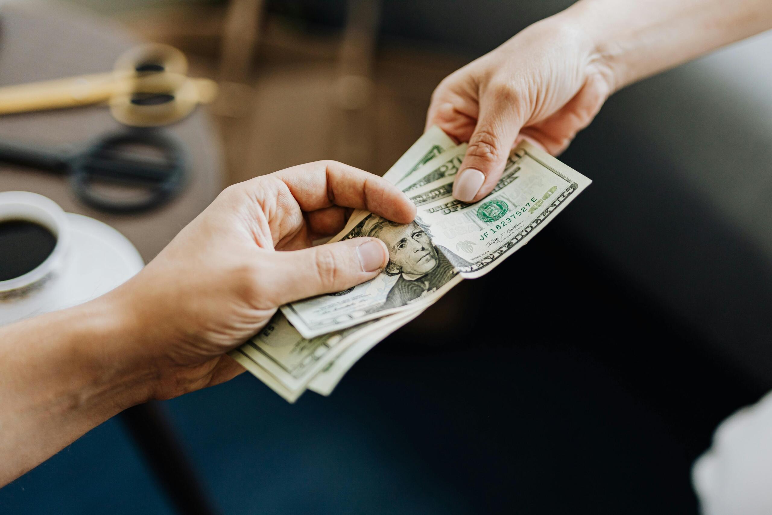 Two hands exchanging a stack of cash, with a cup of coffee on a table in the foreground and scissors in the background.