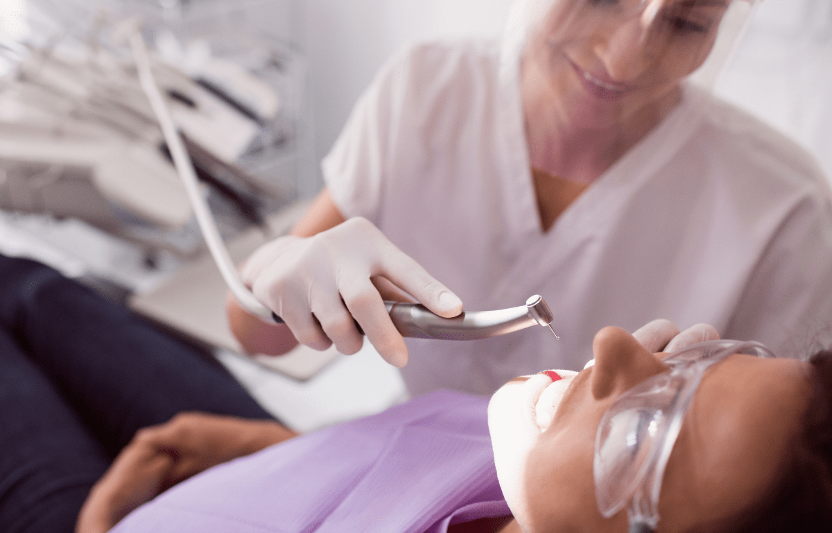A dental professional uses a high-speed handpiece while treating a patient wearing protective eyewear in a bright clinic setting.