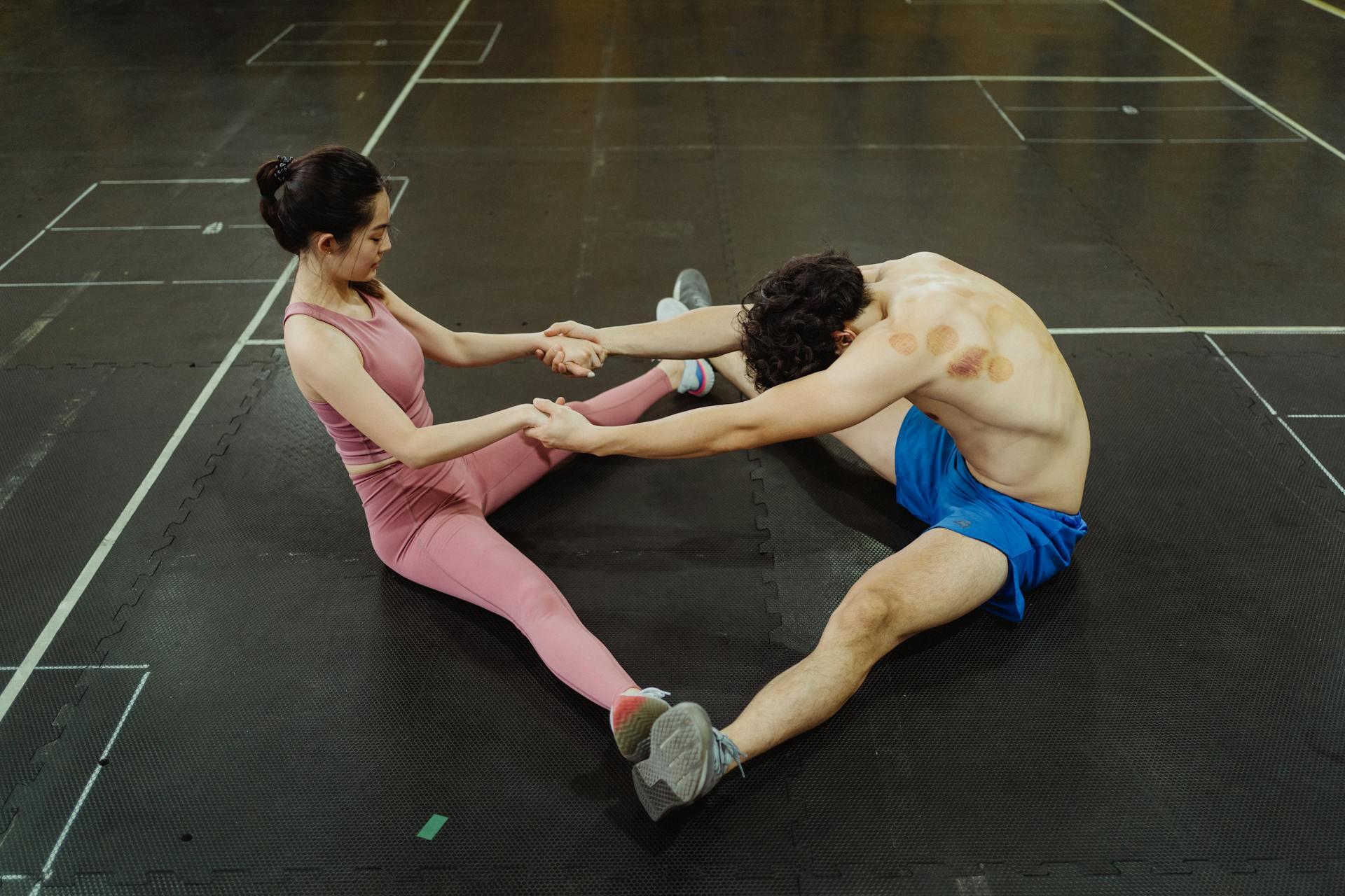 A man and a woman sit on a gym mat, stretching and holding hands, engaged in a warm-up exercise together.