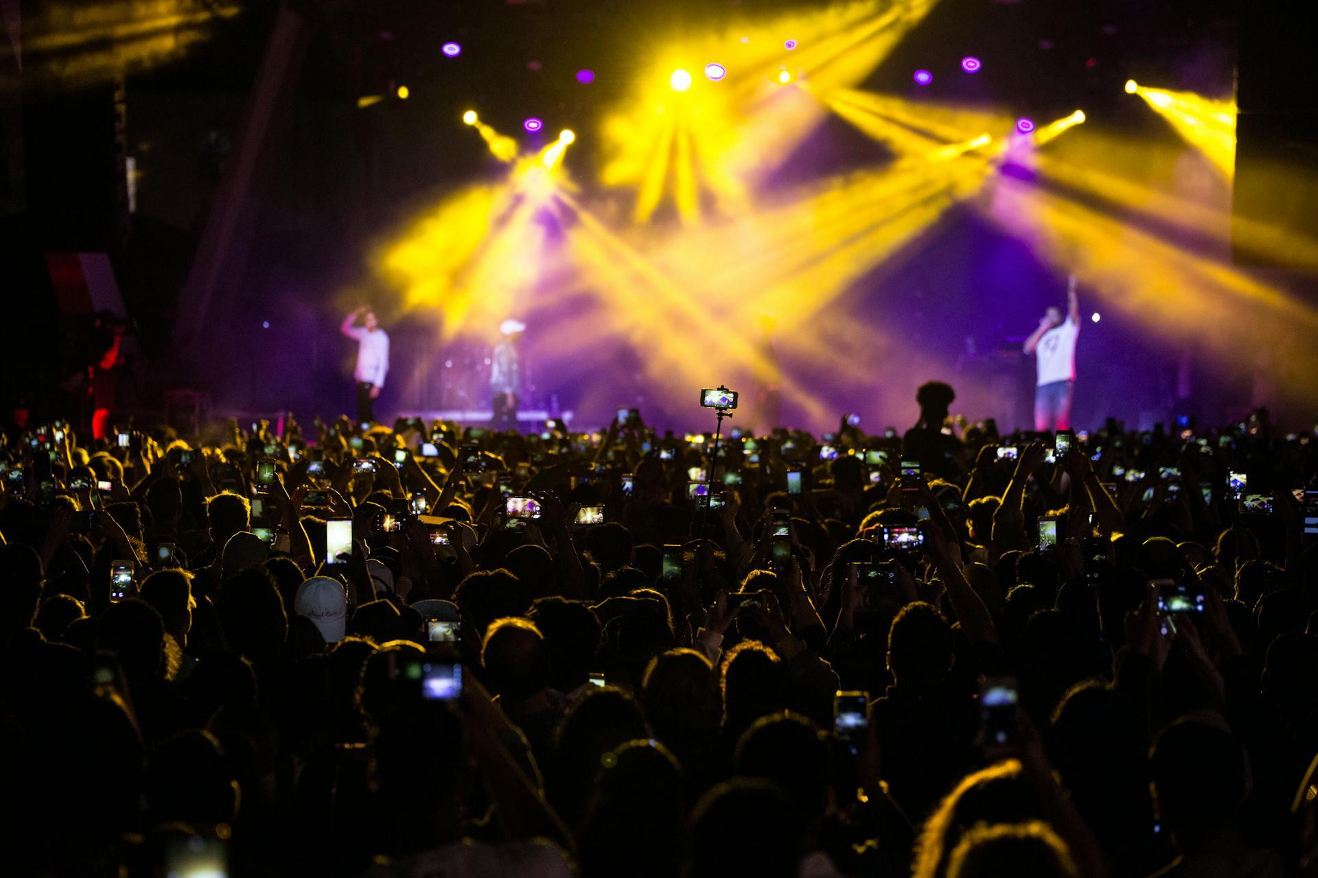 A vibrant concert scene with a large crowd holding smartphones, illuminated by colorful stage lights and two performers in the background.
