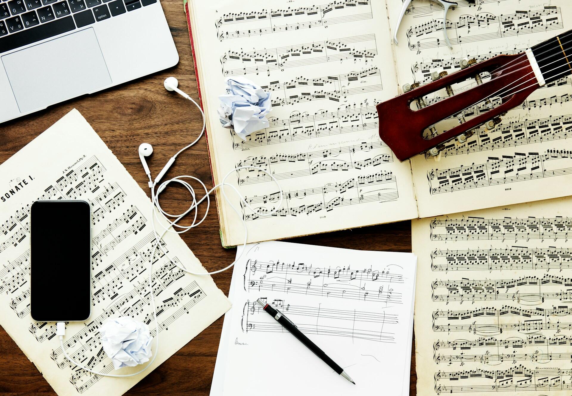 A creative workspace featuring sheet music, a smartphone, headphones, a sketch of music notes, and a guitar on a wooden table.