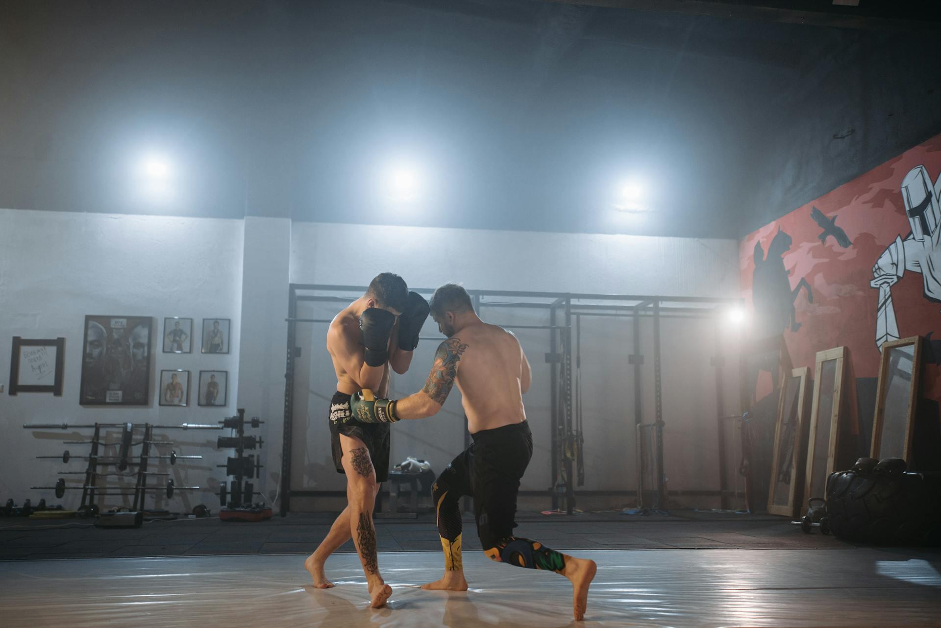 Two shirtless athletes sparring in a gym, with training gear and weights in the background. Bright lighting enhances the action.