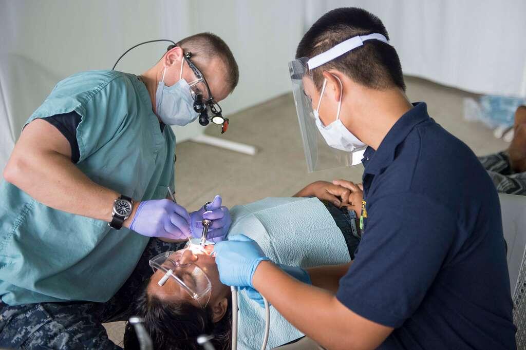 A dental professional examines a patient's mouth while an assistant holds tools and provides support during the procedure.