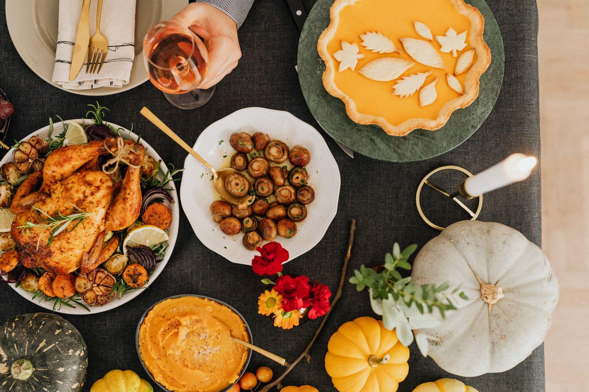 An overhead table holds roast chicken, sautéed mushrooms, pumpkin pie, an orange side dish, pumpkins, flowers, and a hand pouring a drink.