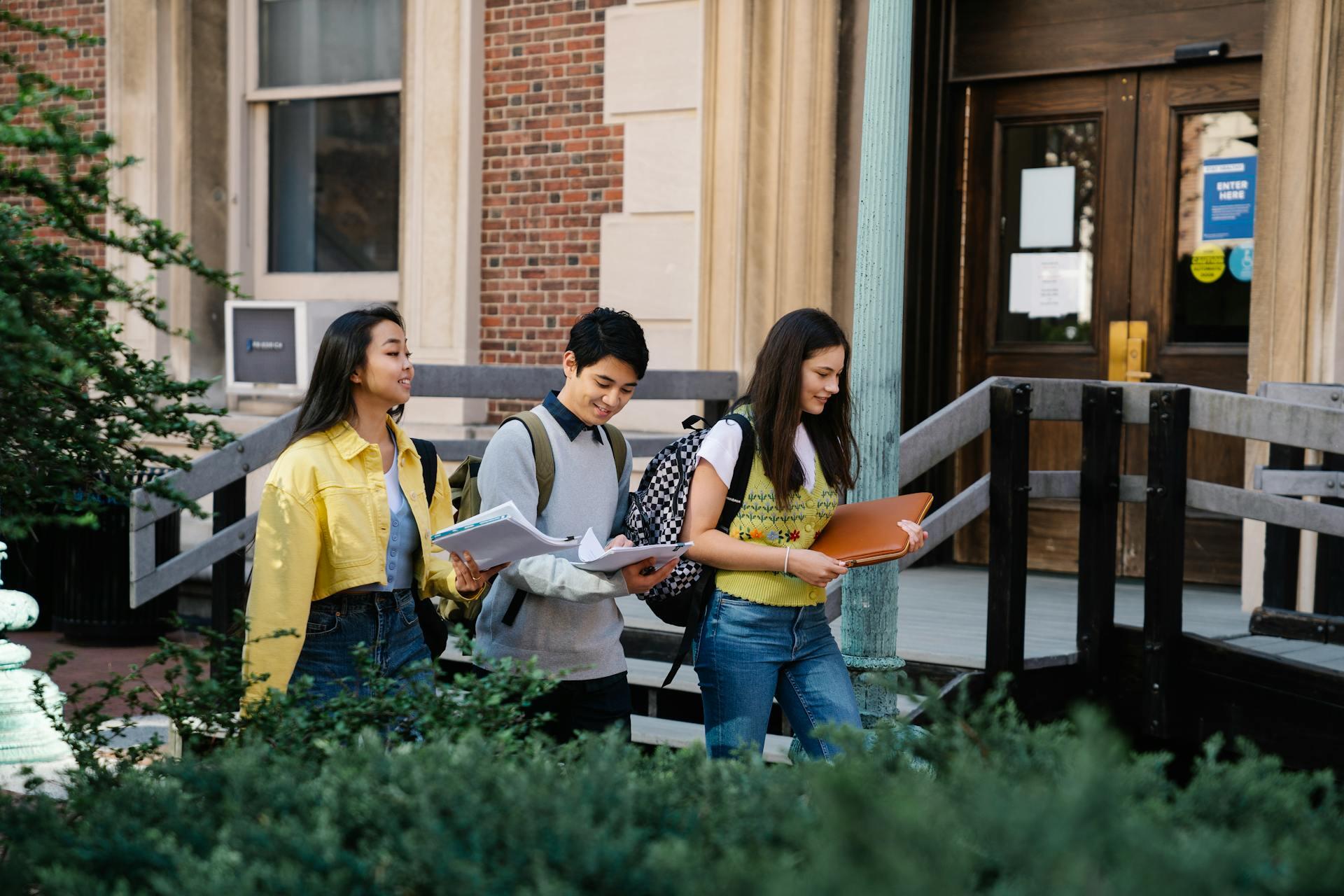 Three students walk together outside a brick building, holding notebooks and a laptop, surrounded by greenery.