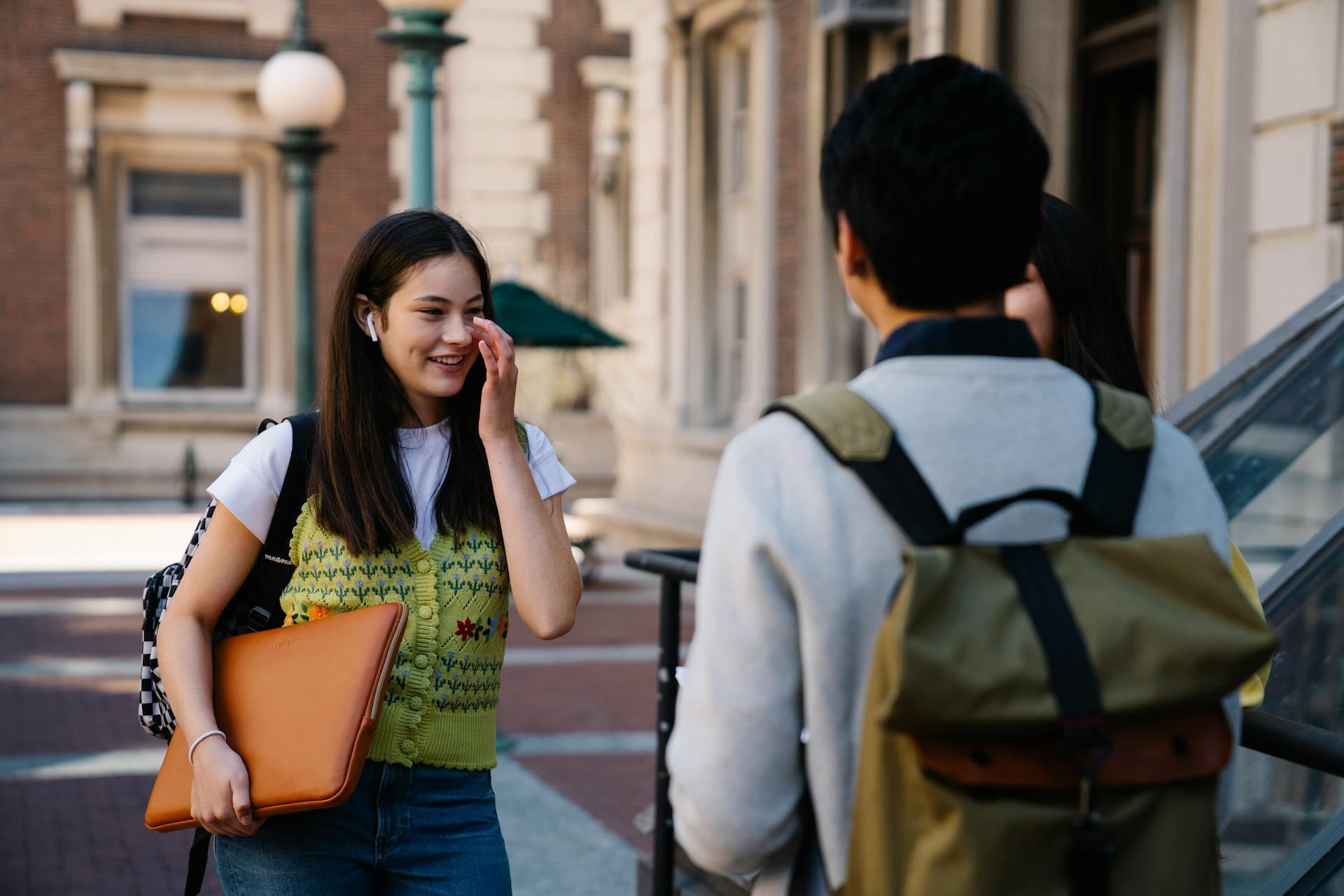 A young woman wearing a green floral sweater and holding a laptop sleeve stands talking with a man in a gray sweater and backpack.