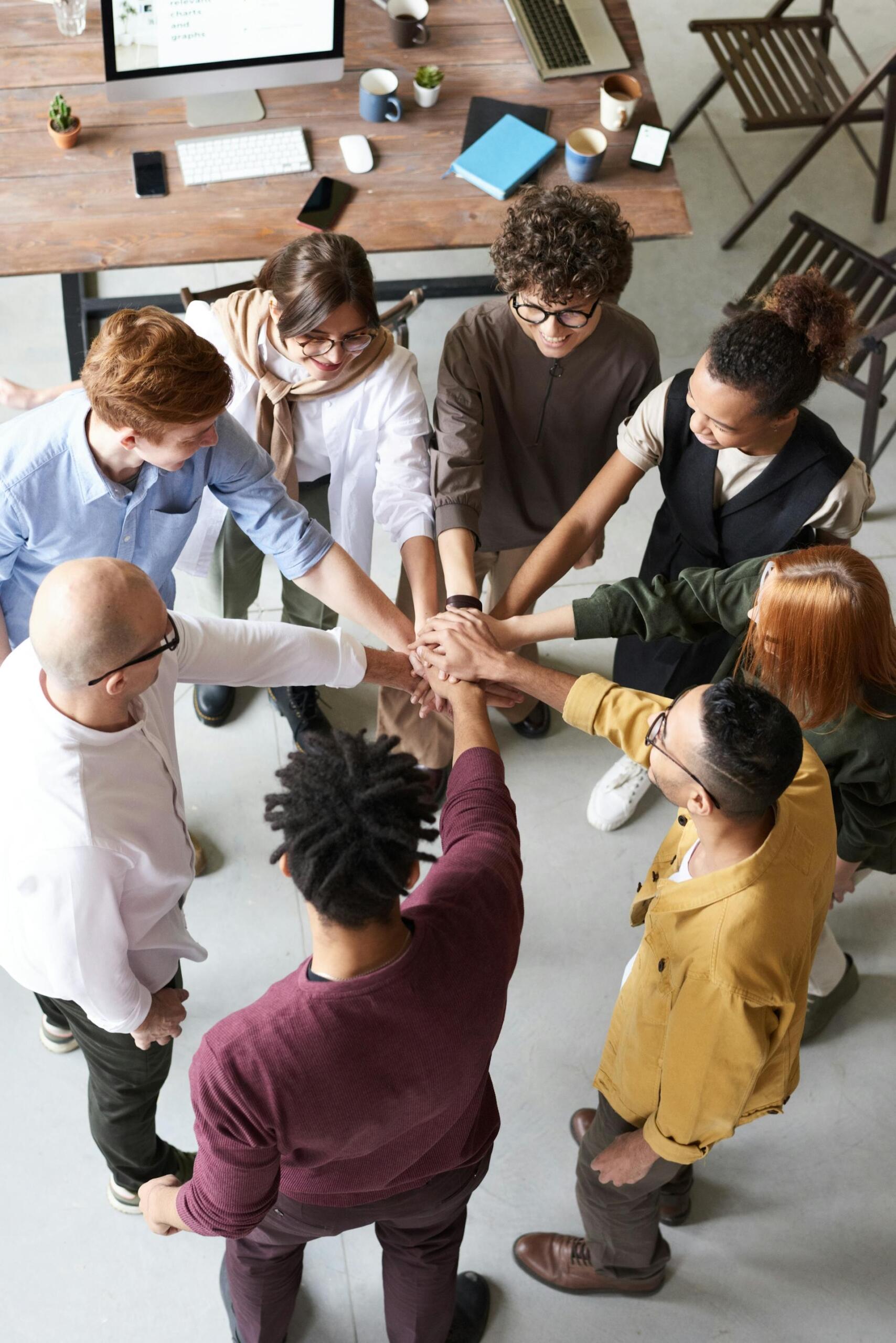 A diverse group of people join hands in a motivational team gesture, surrounded by office supplies and a computer on a wooden table.