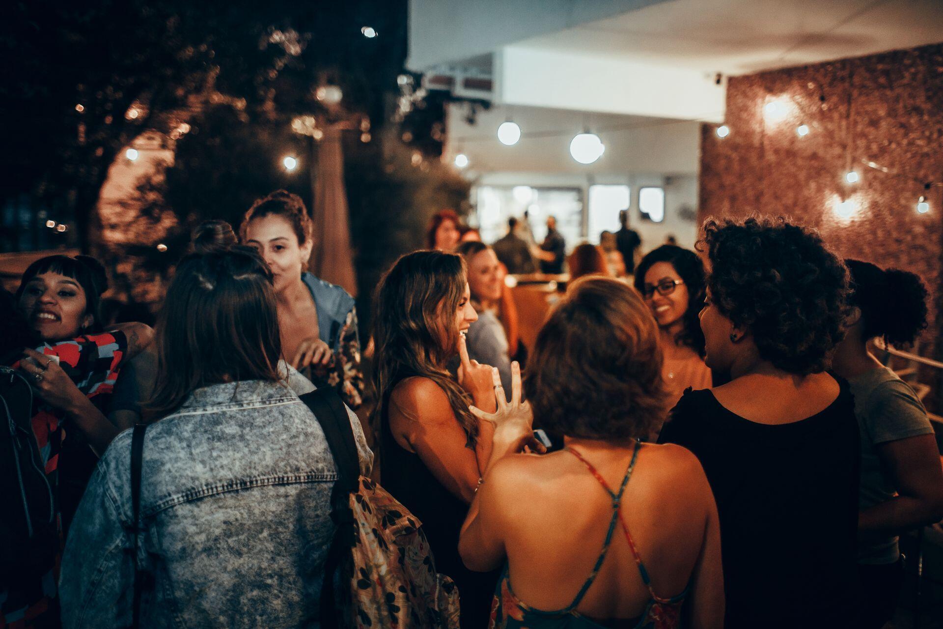A crowd of people talk in small groups at a warmly lit indoor gathering, with several people seated in the background.
