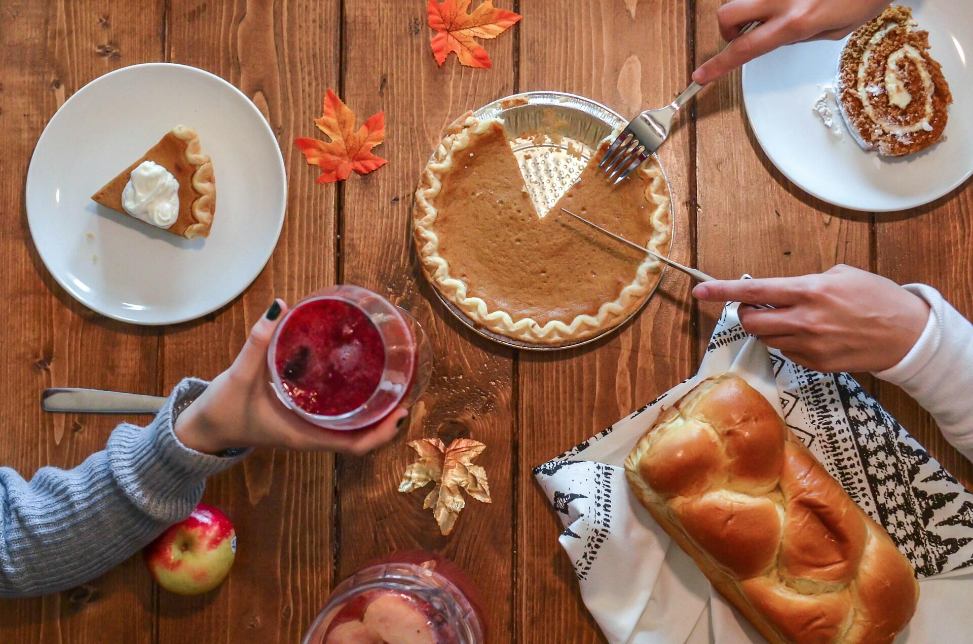 A wooden table set for Thanksgiving, adorned with autumn leaves features pumpkin pie, a glass of berry drink, a loaf of bread, and a slice of pumpkin roll cake.