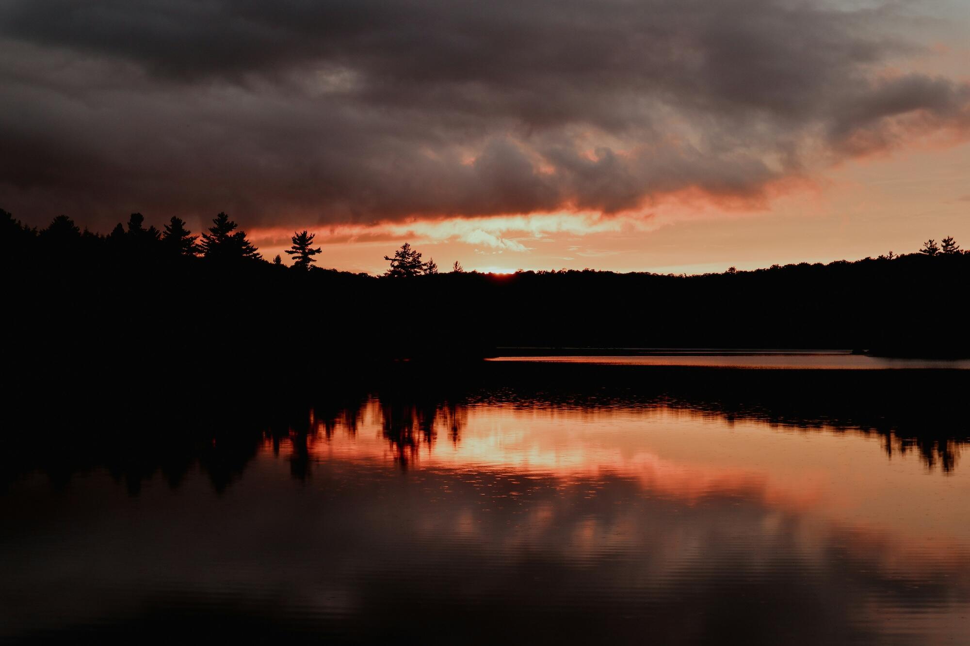 The sun sets over a calm lake, painting the sky with warm colors, while trees stand quietly along the water's edge.