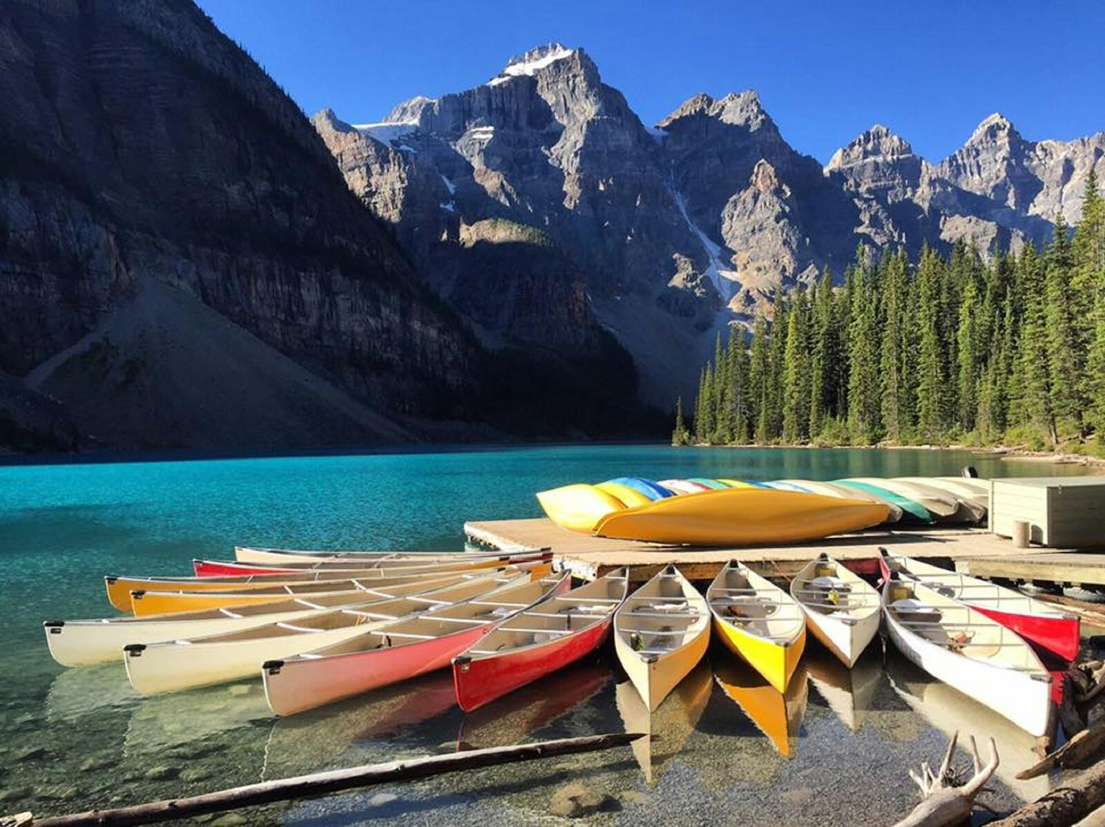 Colorful canoes lined up on a dock by a turquoise lake, surrounded by towering mountains and lush green trees under a clear blue sky.
