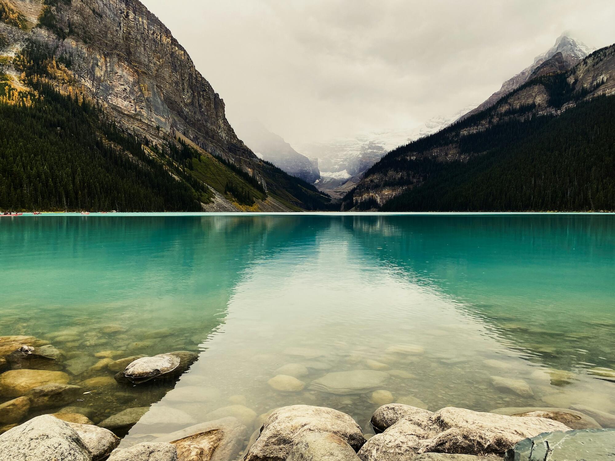 A tranquil turquoise lake surrounded by towering mountains and lush forests, reflecting a cloudy sky. Smooth stones line the shoreline.