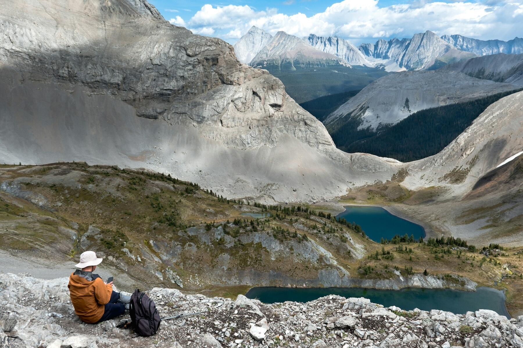 A hiker sits on a rocky ledge, overlooking deep valleys and turquoise lakes, surrounded by rugged mountains under a blue sky.