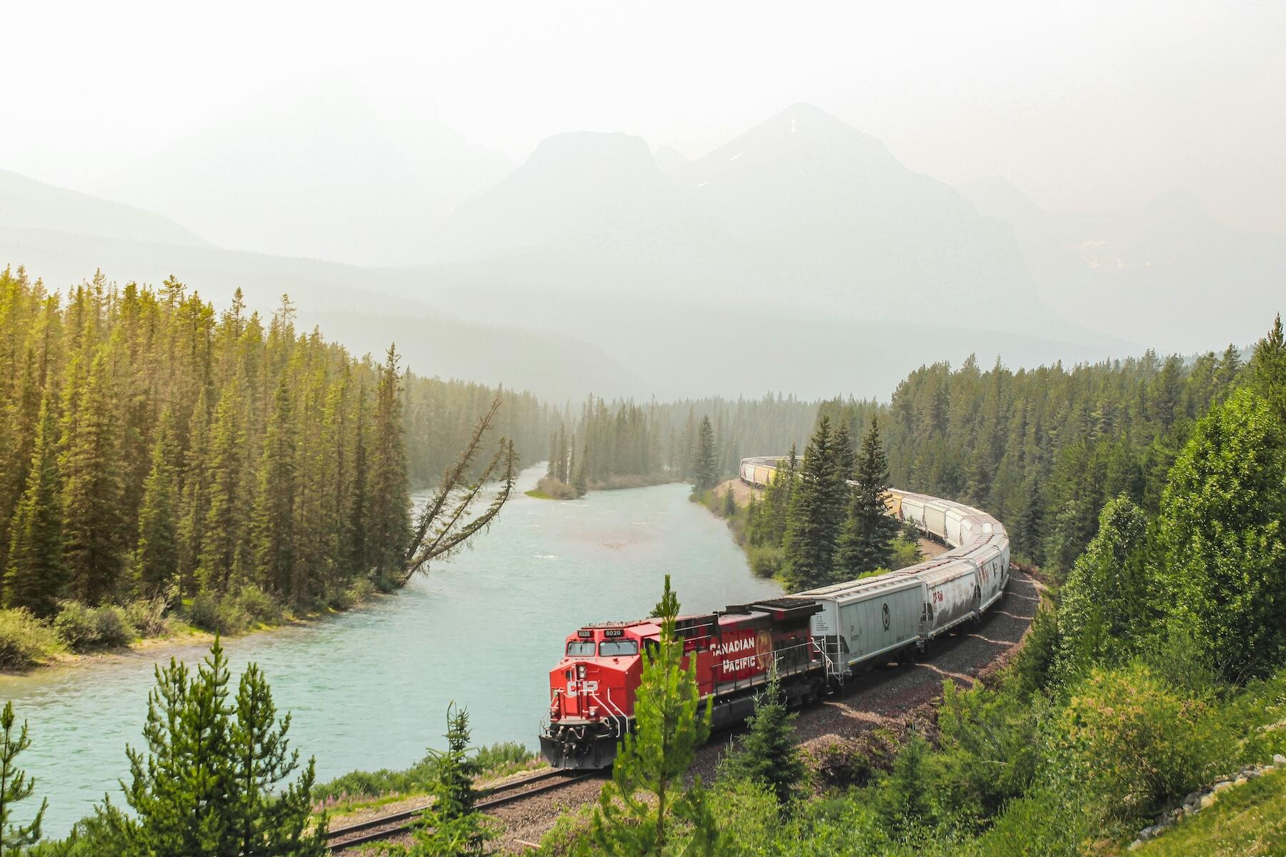 A Canadian Pacific train curving along a river, surrounded by dense evergreen forests and hazy mountains in the background.