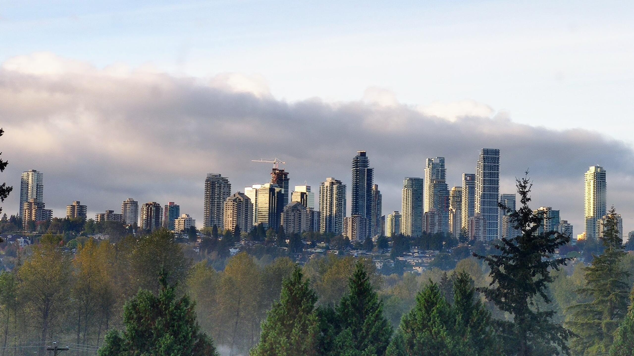 Skyline view of tall modern buildings against a backdrop of clouds, with green trees in the foreground.