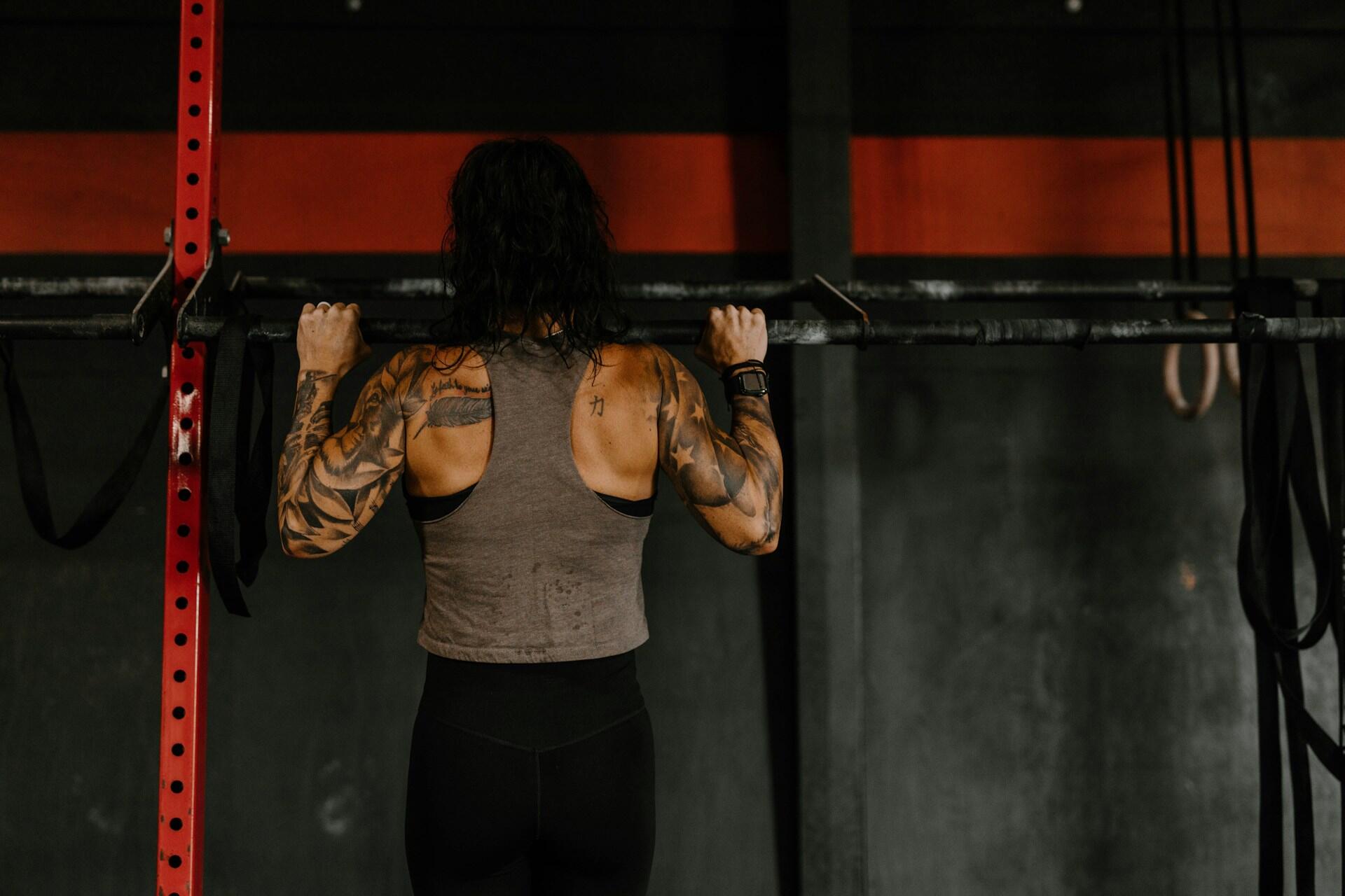 A person with tattoos performs a pull-up on a bar in a gym, showcasing strength and focus against a dark background.