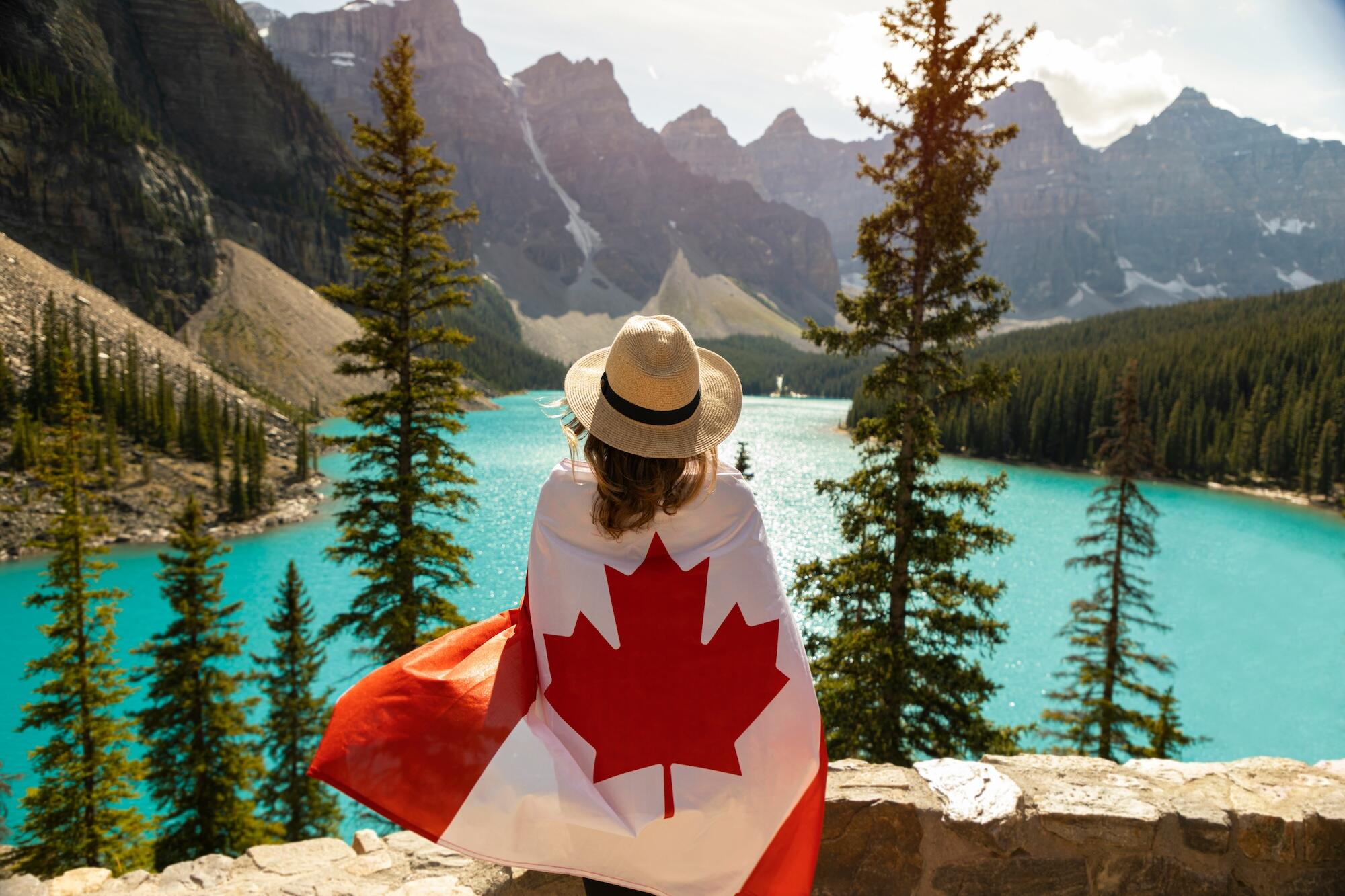 A person in a straw hat wraps themselves in a Canadian flag, overlooking a turquoise lake surrounded by mountains and pine trees.