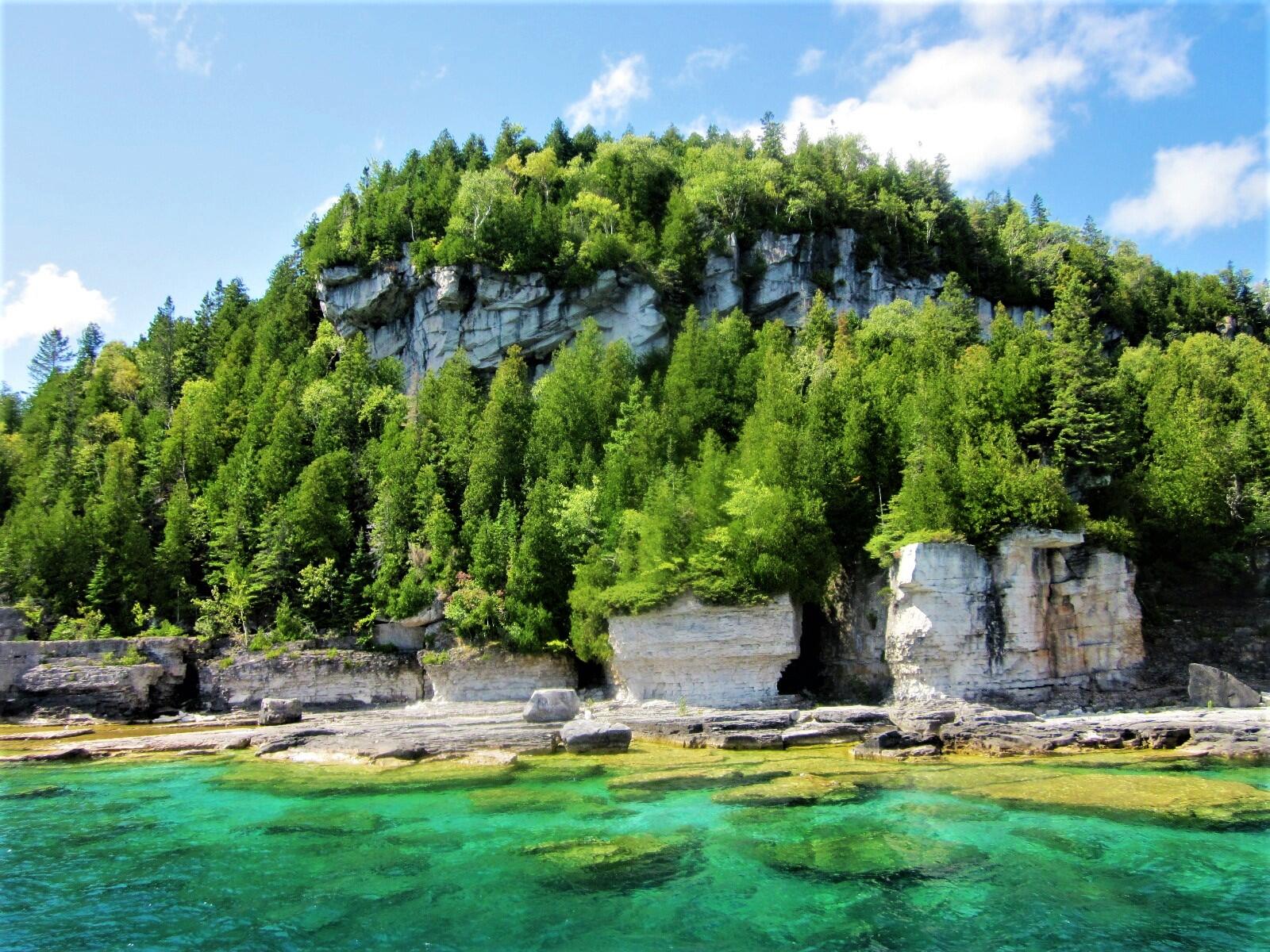 Lush green cliffs rise above clear turquoise waters, with rocky shores and trees dotting the landscape under a bright blue sky.