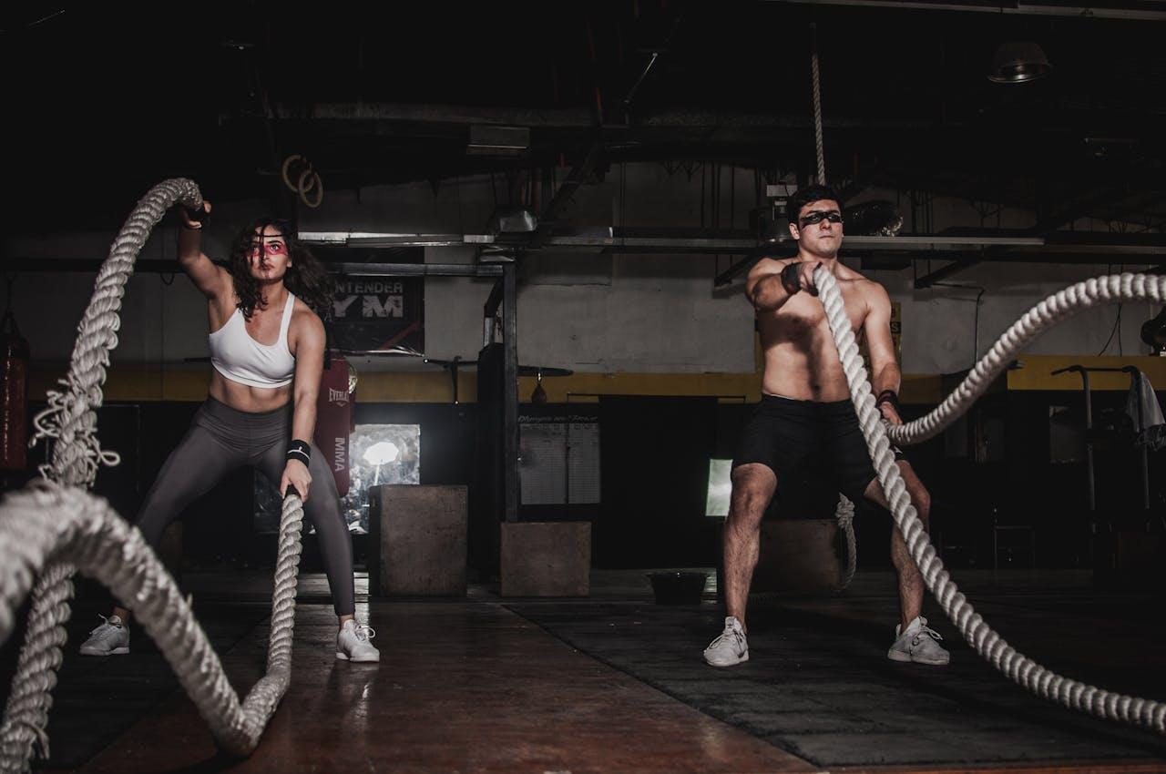 Two individuals perform battle rope exercises in a dimly lit gym, showcasing their strength and focus during a workout session.