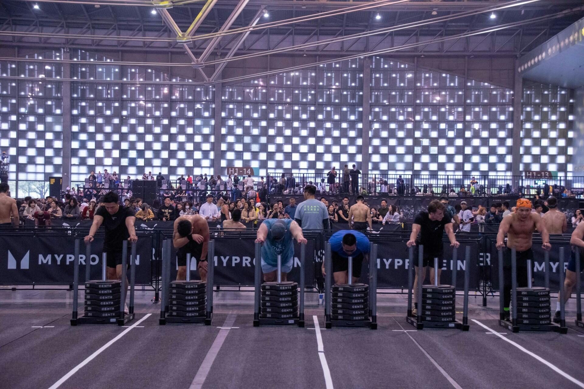 Athletes compete in a fitness event, pushing sleds on a track, while a crowd cheers in a large indoor arena.