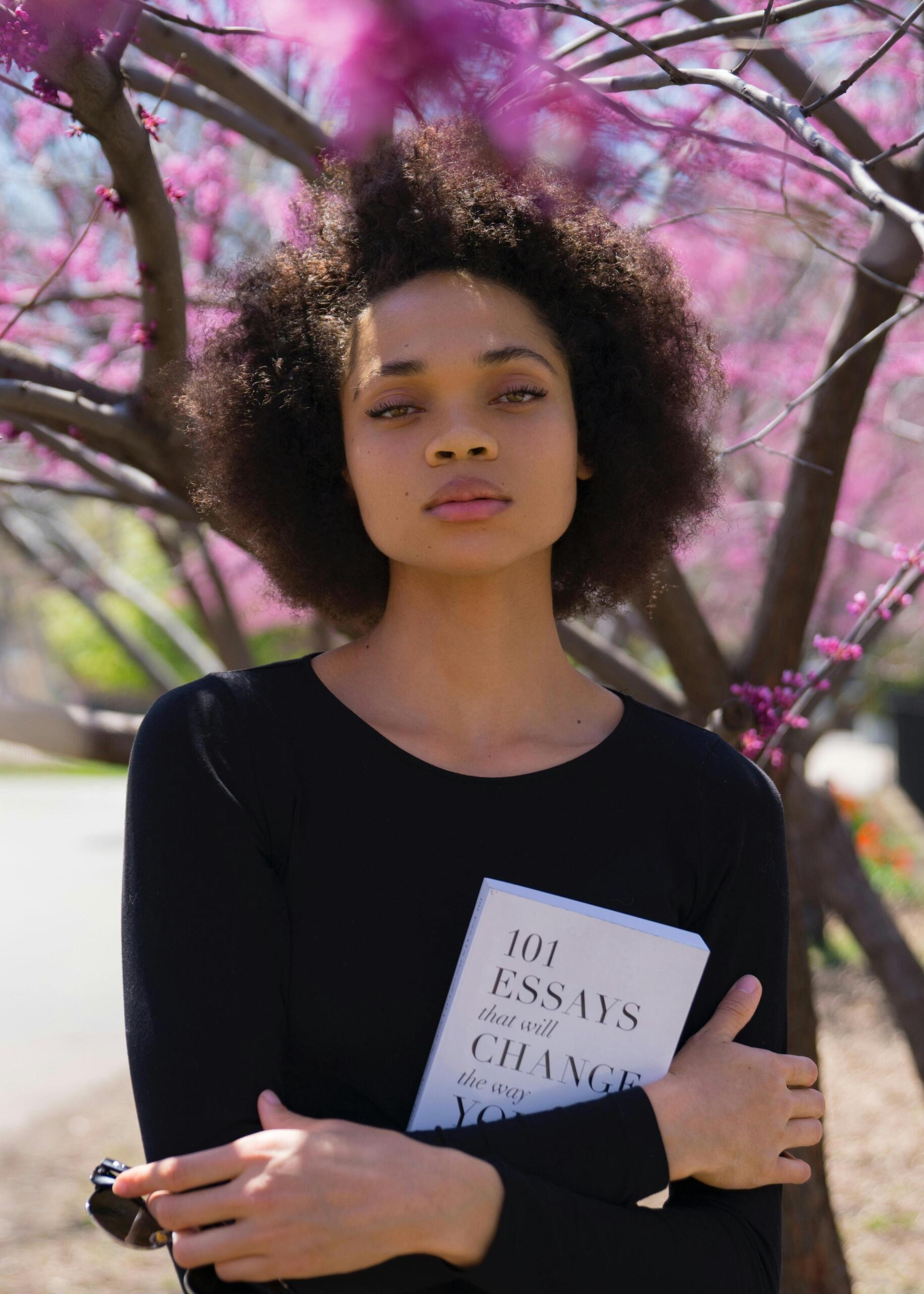 A woman holds a book and looks toward the camera while standing in front of blossoming trees, with soft spring flowers in the background. Source: Thought Catalog.