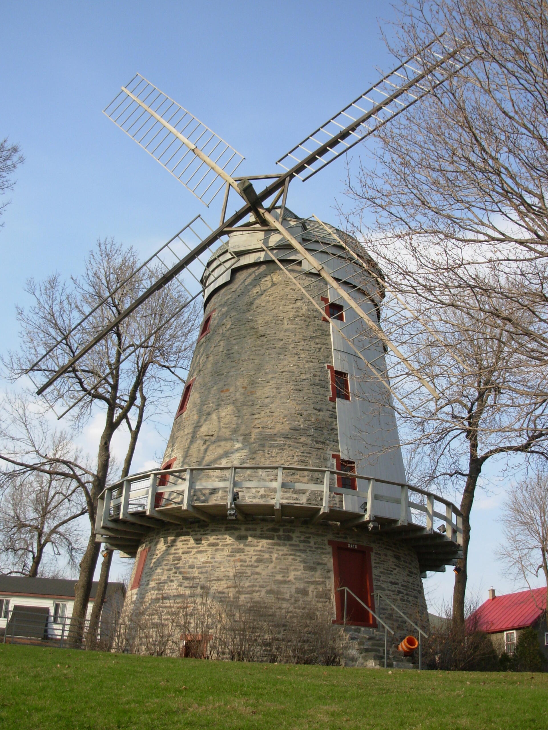 A historic stone windmill with a wooden balcony, surrounded by grass and trees, against a clear blue sky.