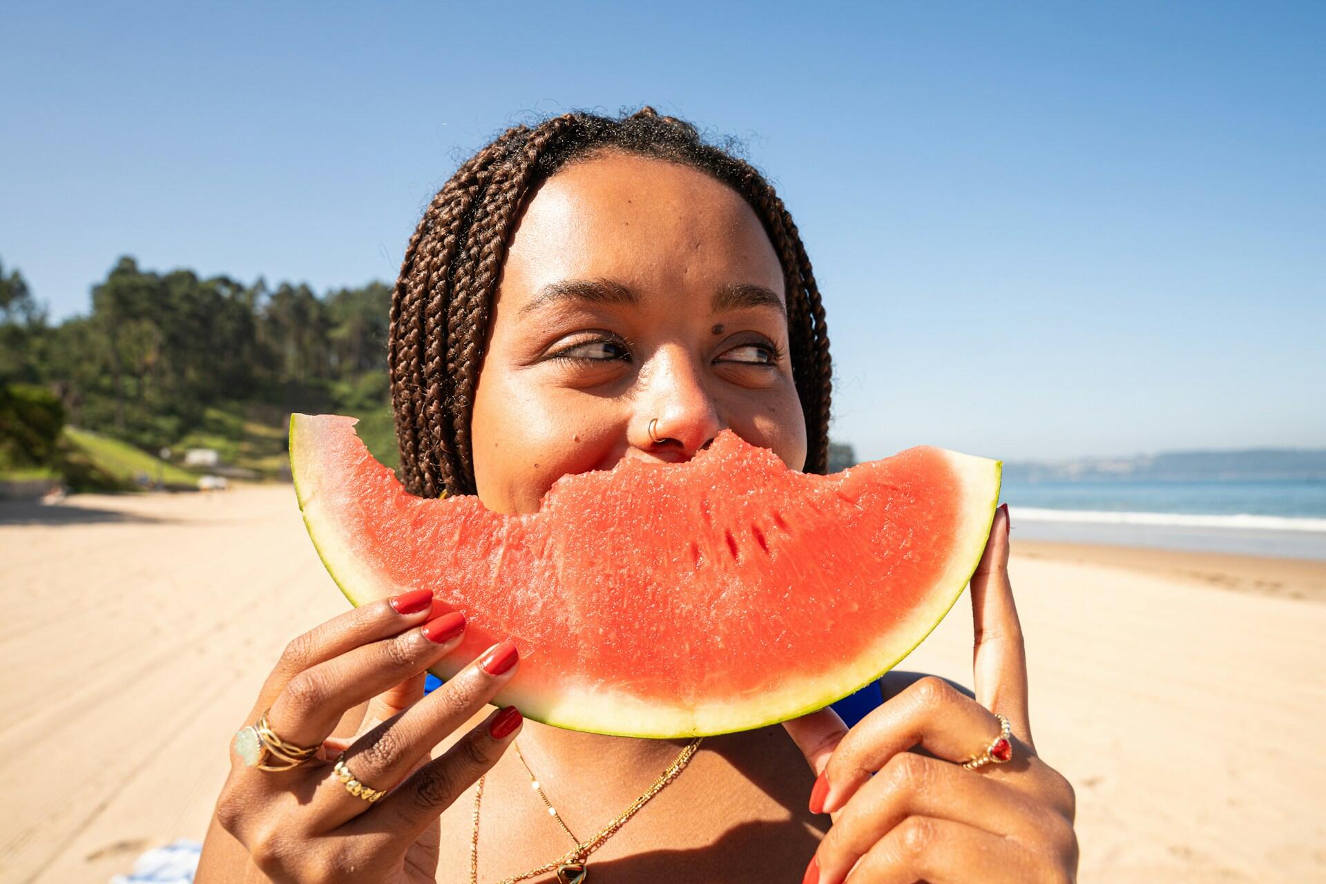 A person holds a slice of watermelon in front of them, with a beach and ocean visible in the background under a clear blue sky.