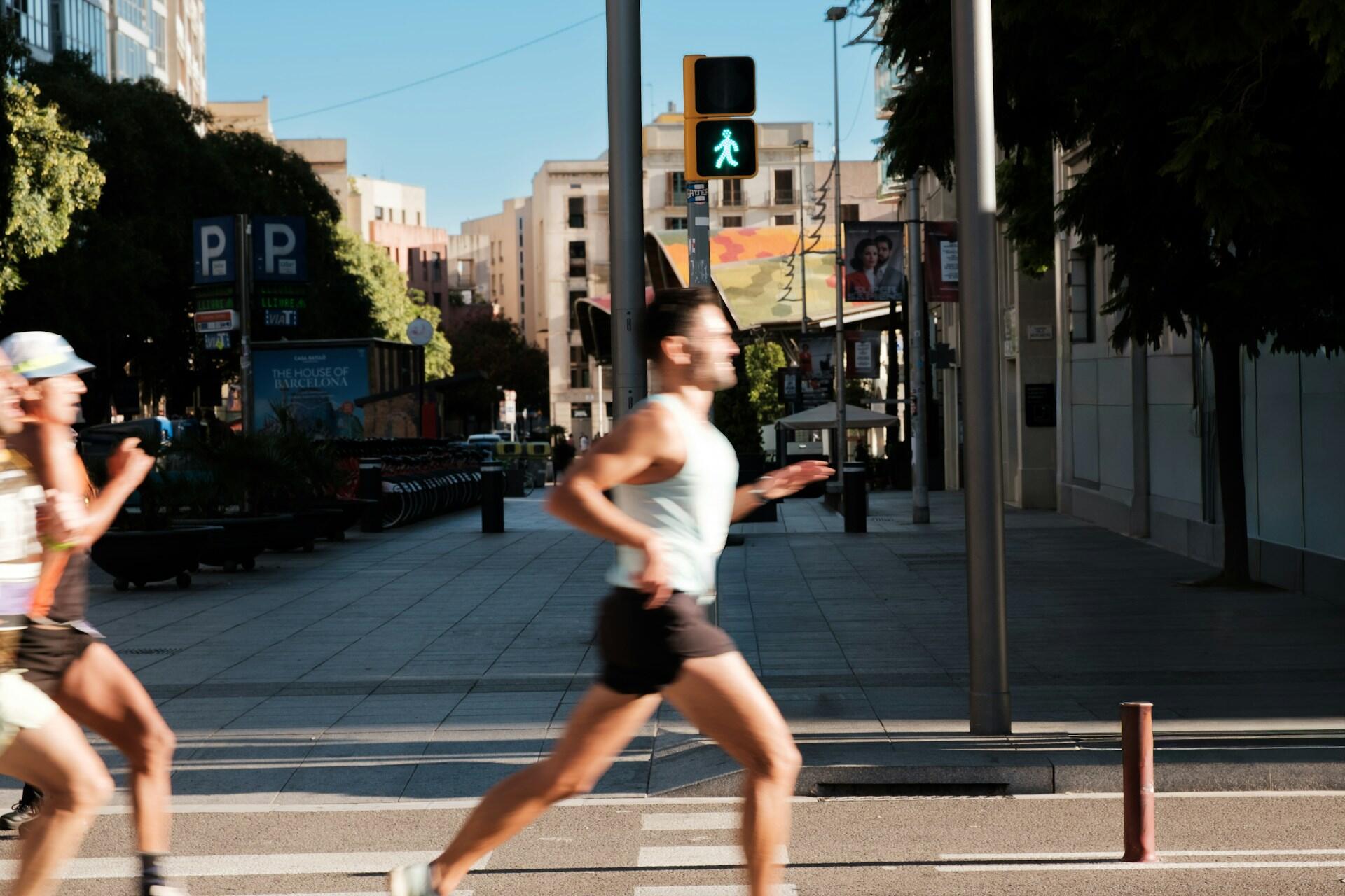 Two runners sprint across a city street with a green pedestrian signal, surrounded by urban scenery and trees in bright daylight.