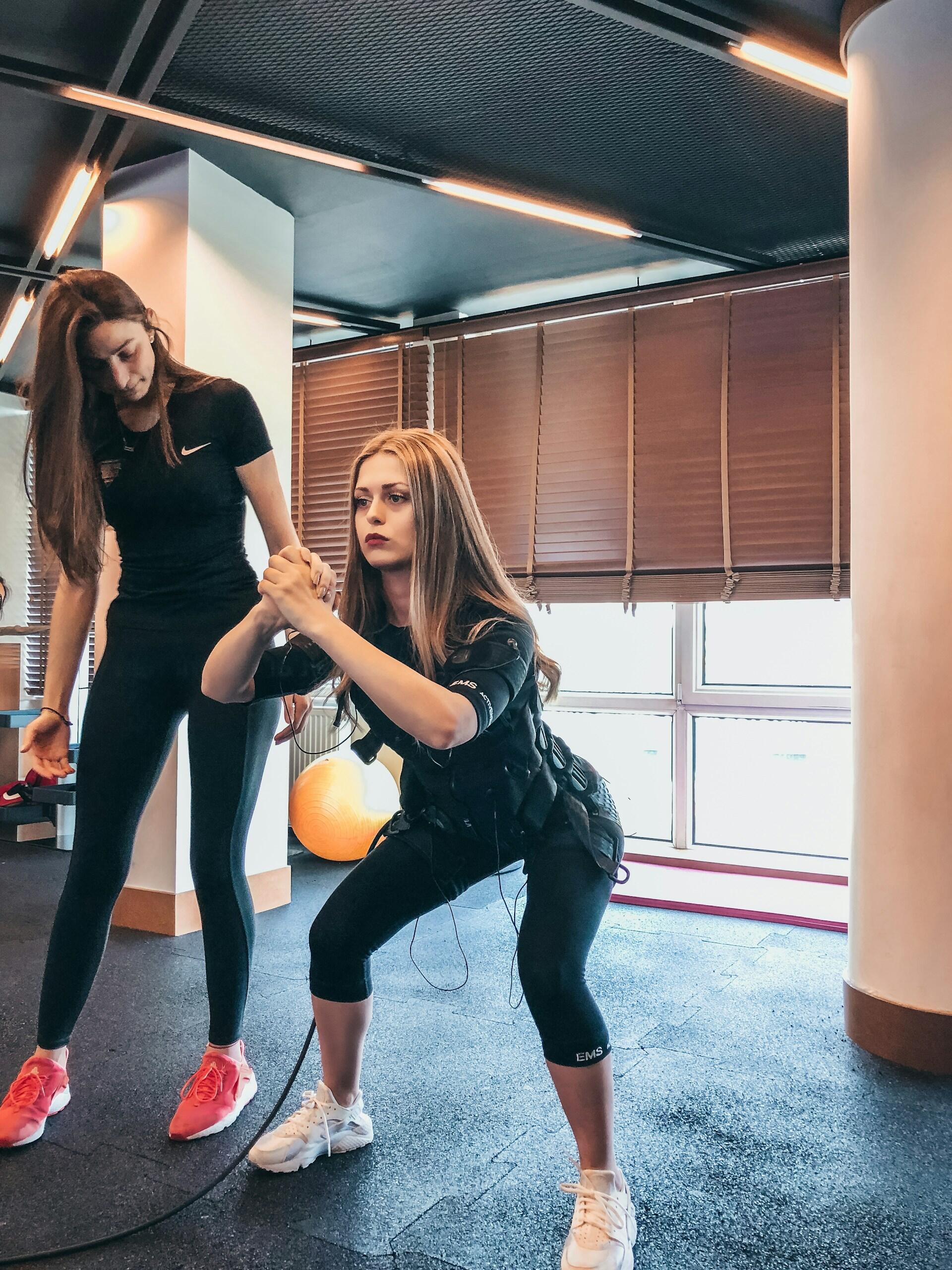 A woman in fitness attire performs a squat while wearing an EMS suit, with another person supporting her in a gym setting.