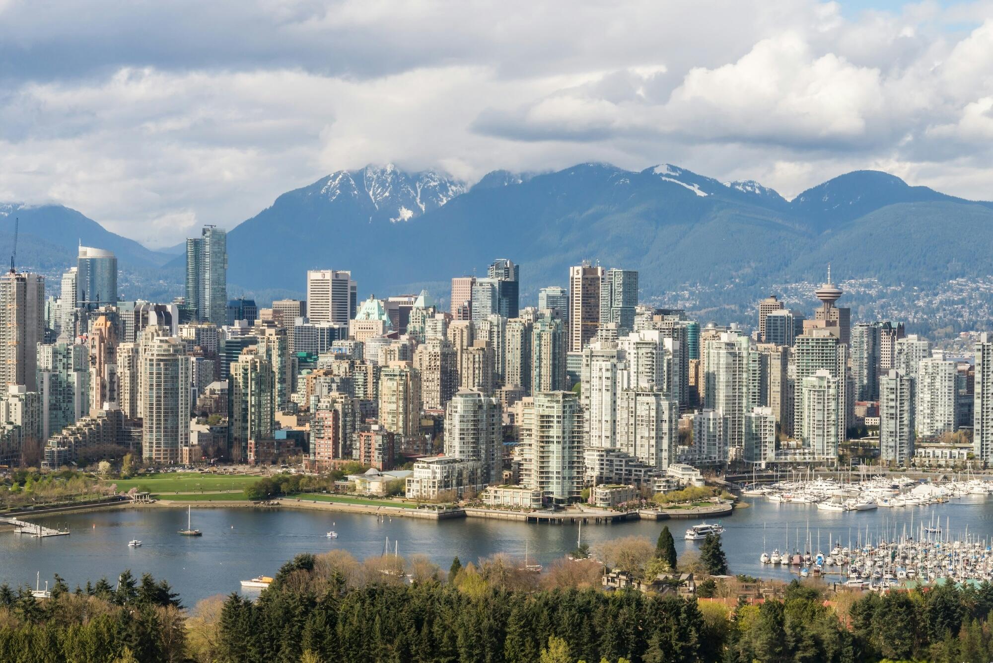 A panoramic view of Vancouver's skyline with skyscrapers, a marina, and snow-capped mountains in the background under a partly cloudy sky.