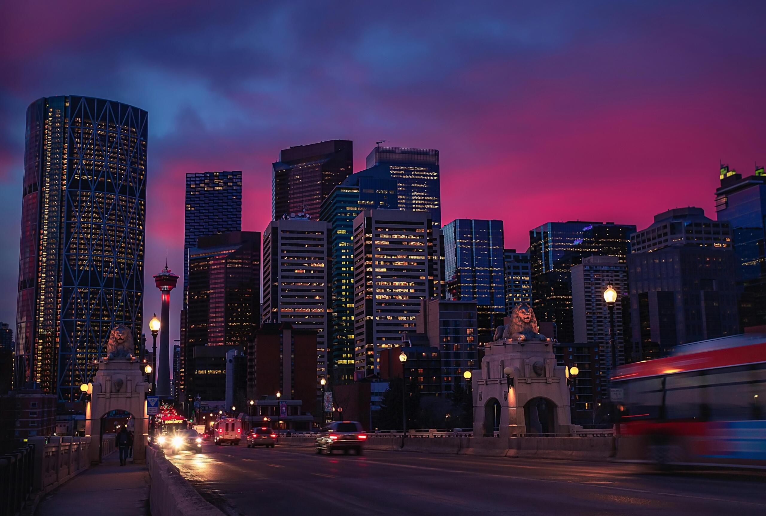 Calgary skyline at dusk featuring modern skyscrapers, a historic bridge with lion statues, and vibrant purple clouds.