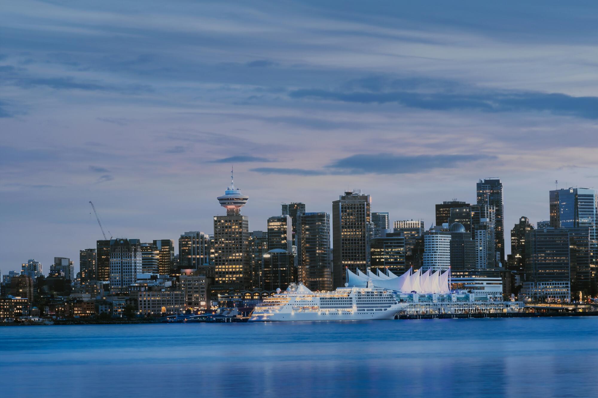 City skyline at dusk featuring tall buildings and a large cruise ship docked by the waterfront, with a soft blue sky backdrop.