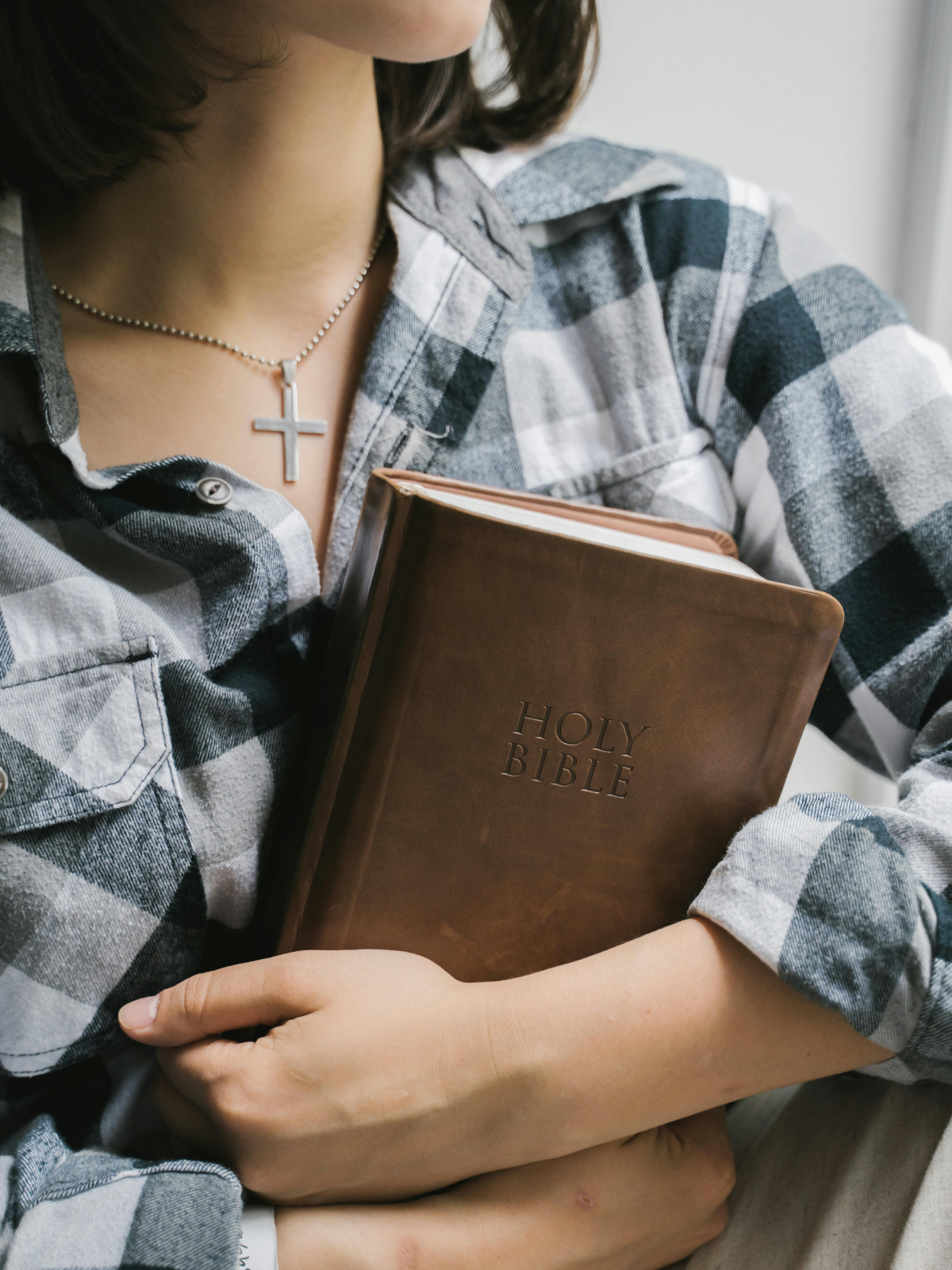 A person in a black and white plaid shirt holds a brown leather-bound Bible close to their chest, wearing a silver cross necklace.