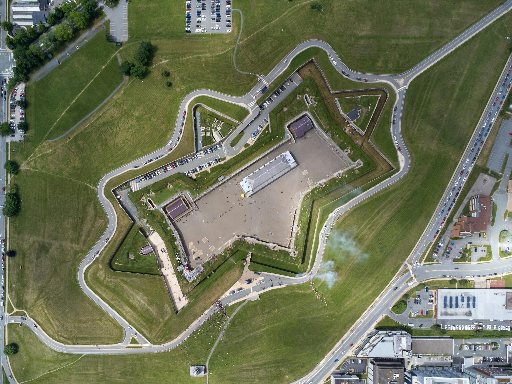 Aerial view of a star-shaped fort surrounded by green lawns and winding roads, with parked cars and people in the courtyard below.