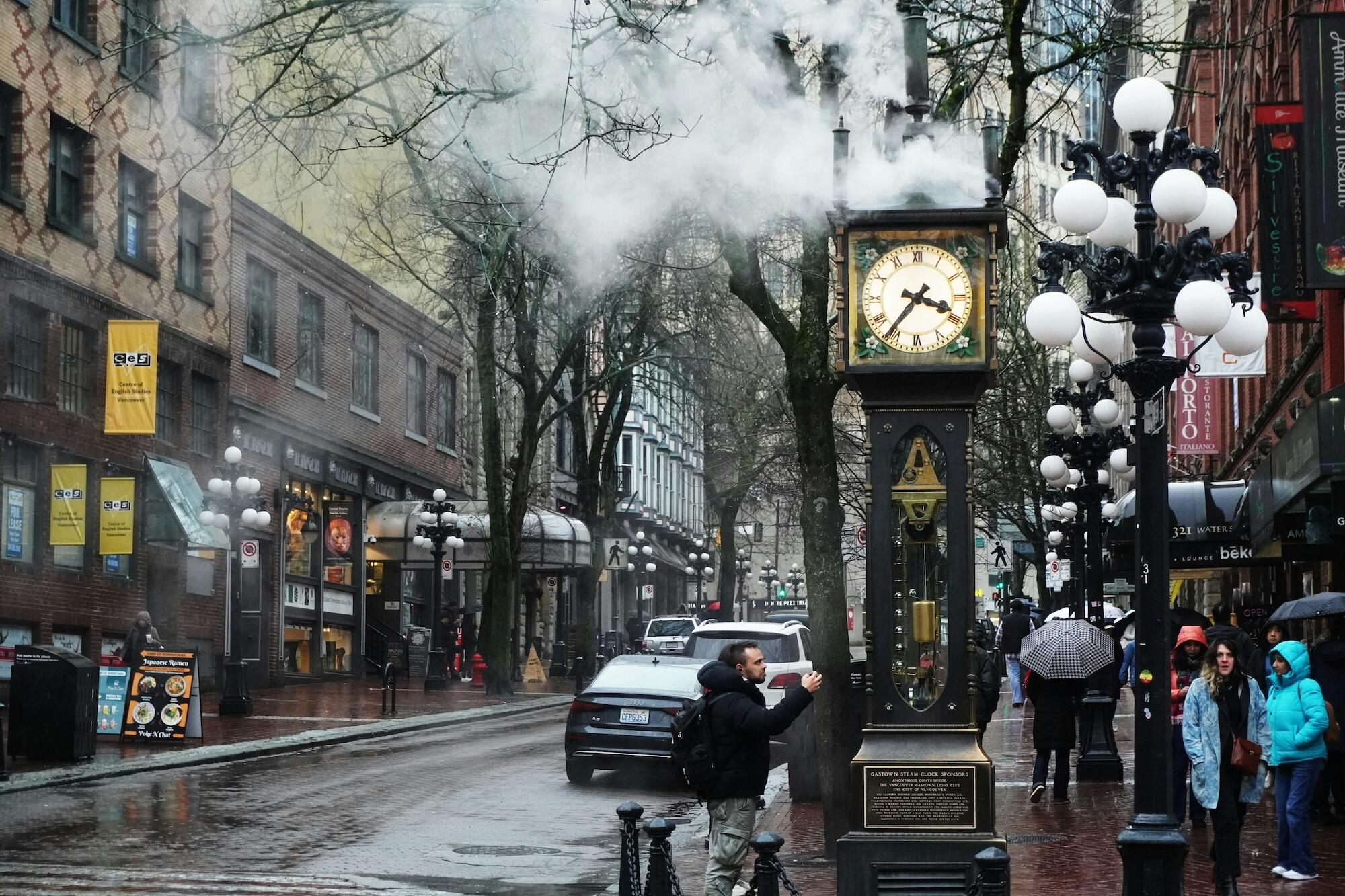 A vintage steam clock stands on a rainy street, surrounded by shops and trees, with steam billowing from its top.