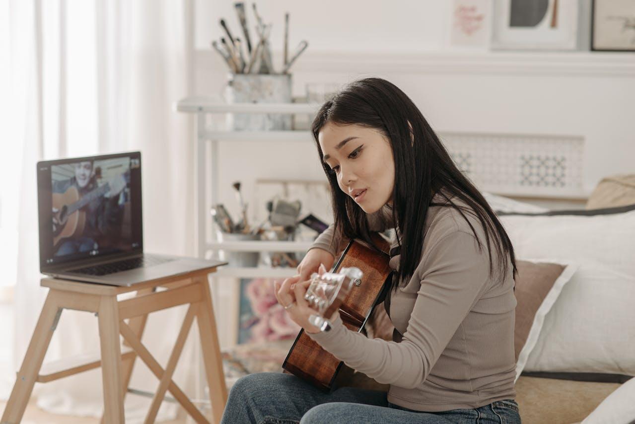 A person sits on a bed, playing guitar while watching a video lesson on a laptop set on a wooden stand. The room is cozy and well-decorated.