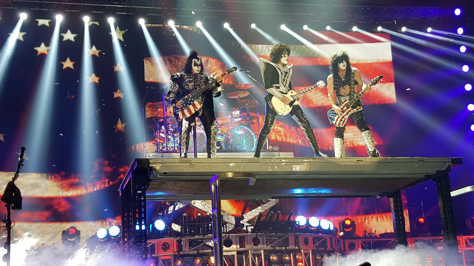 Three musicians perform on stage with electric guitars, surrounded by bright lights and a backdrop of American flags and stars.