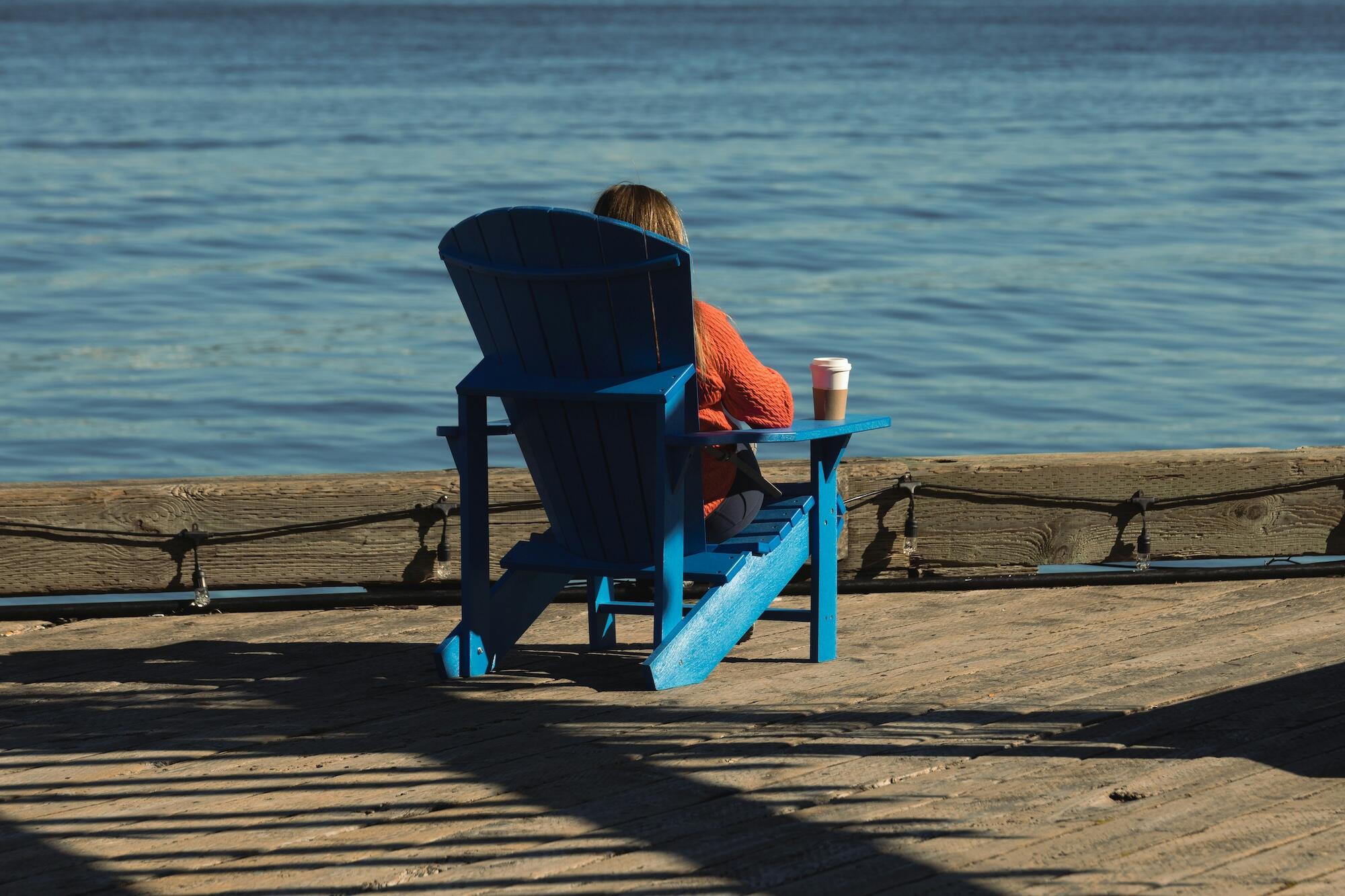 A person in an orange sweater relaxes in a blue chair by the water, holding a coffee cup, enjoying a serene view.