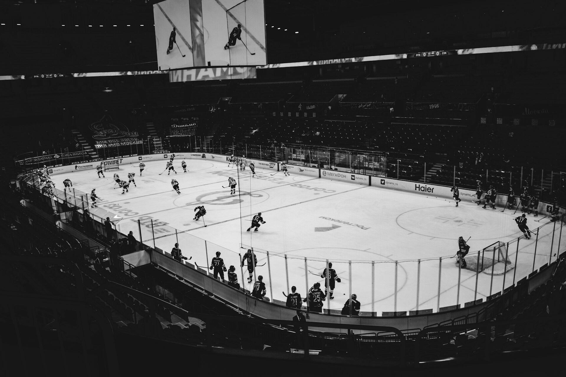 A black and white image of an ice hockey rink with players warming up, surrounded by empty stands and large screens above.