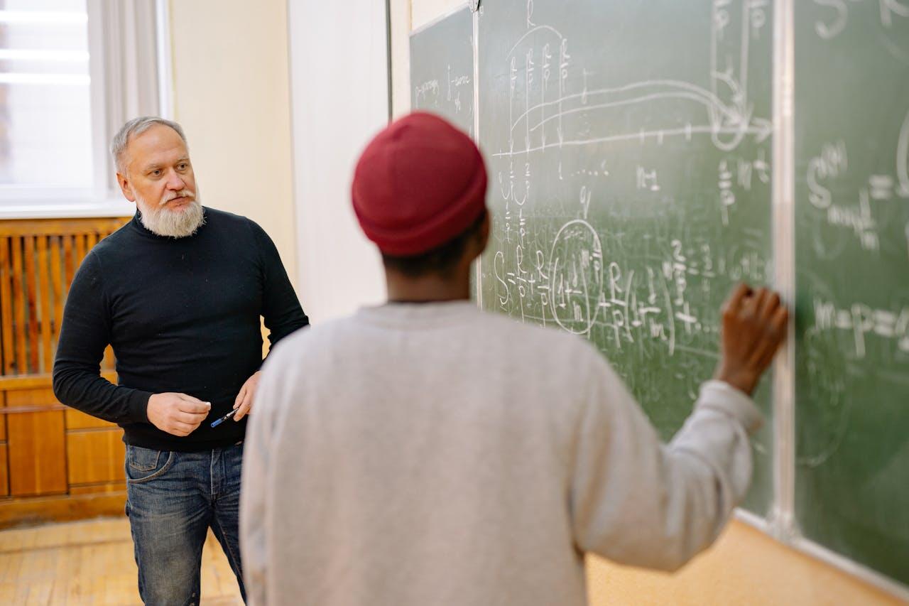 A student in a gray sweatshirt writes on a chalkboard filled with mathematical equations while an instructor observes.