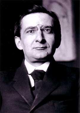 A man in a formal black suit and tie, seated against a dark background, with a vintage style. 